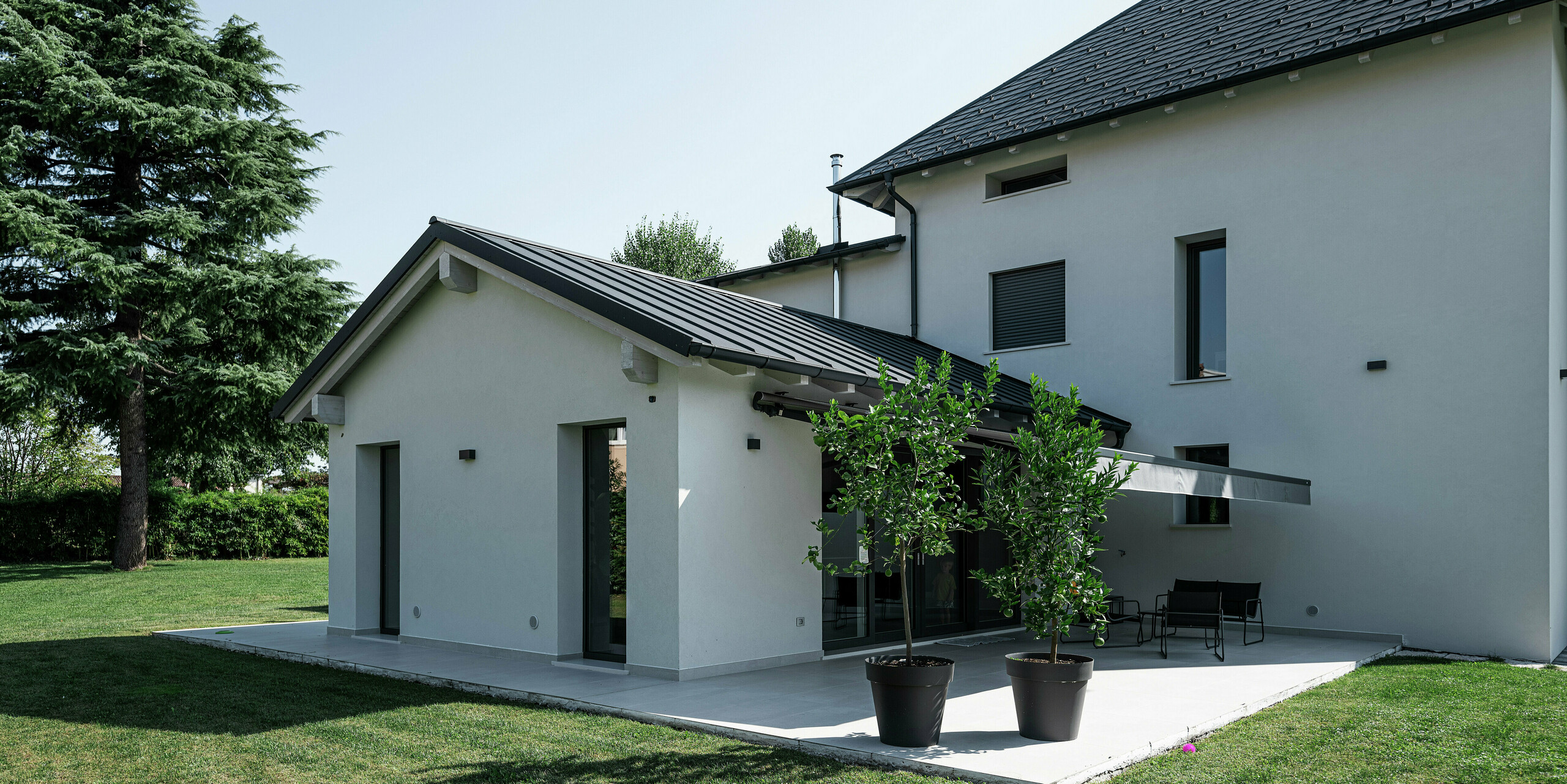 Nahaufnahme des Anbaus eines modernen Einfamilienhauses in Marano Vicentino, Italien, mit einer PREFA Doppelstehfalzdeckung in P.10 Anthrazit. Das elegante Aluminiumdach ergänzt harmonisch die helle Fassade und sorgt für eine langlebige und witterungsbeständige Lösung. Die angrenzende Terrasse mit Sonnenschutz schafft einen angenehmen Außenraum im Garten.