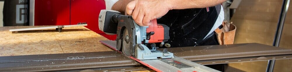 Das Bild zeigt einen Mann, der an einer Werkbank arbeitet. Er schneidet mit einer Sägemaschine das PREFA Siding-Fassadenelement zu. Er ist dabei sehr fokussiert, die Detailgenauigkeit seiner Arbeit kommt zum Vorschein.