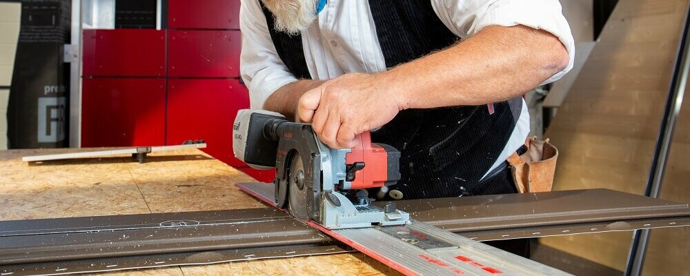 Das Bild zeigt einen Mann, der an einer Werkbank arbeitet. Er schneidet mit einer Sägemaschine das PREFA Siding-Fassadenelement zu. Er ist dabei sehr fokussiert, die Detailgenauigkeit seiner Arbeit kommt zum Vorschein.