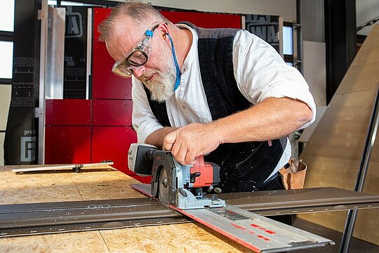 Das Bild zeigt einen Mann, der an einer Werkbank arbeitet. Er schneidet mit einer Sägemaschine das PREFA Siding-Fassadenelement zu. Er ist dabei sehr fokussiert, die Detailgenauigkeit seiner Arbeit kommt zum Vorschein.