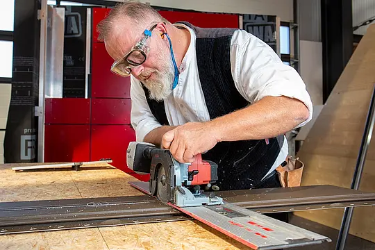 Das Bild zeigt einen Mann, der an einer Werkbank arbeitet. Er schneidet mit einer Sägemaschine das PREFA Siding-Fassadenelement zu. Er ist dabei sehr fokussiert, die Detailgenauigkeit seiner Arbeit kommt zum Vorschein.