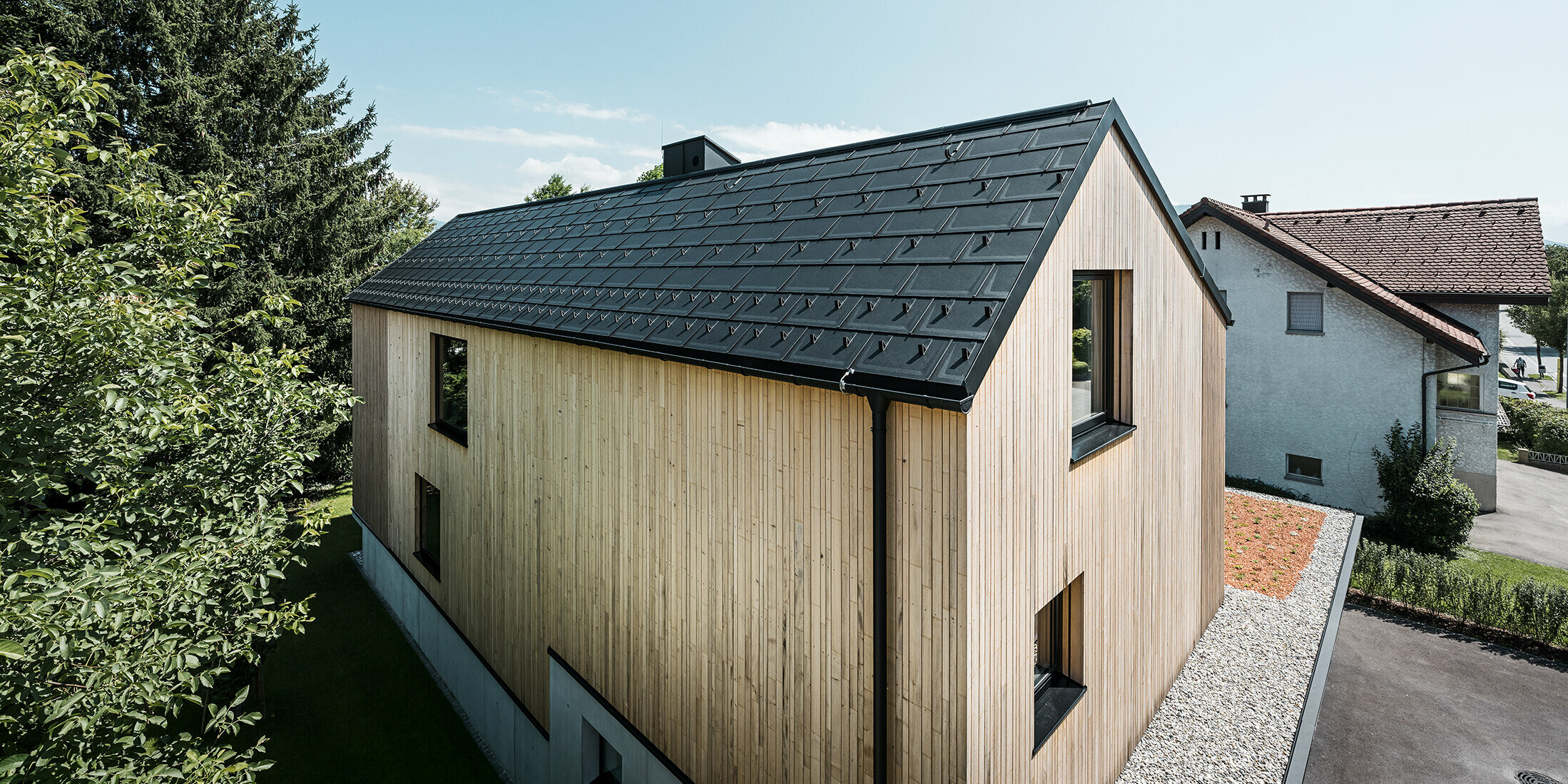 Ein modernes Einfamilienhaus in Lustenau besticht durch die Kombination einer freundlichen Holzfassade mit robusten PREFA Aluminiumprodukten. Das Satteldach wird von der PREFA Dachplatte R.16 mit Schneestoppern sowie der passenden Dachentwässerung in Form von Hängerinne und Ablaufrohr geschmückt. Alle Produkte sind einheitlich im Farbton Schwarz P.10 gehalten und harmonieren perfekt mit der vertikalen Holzfassade.