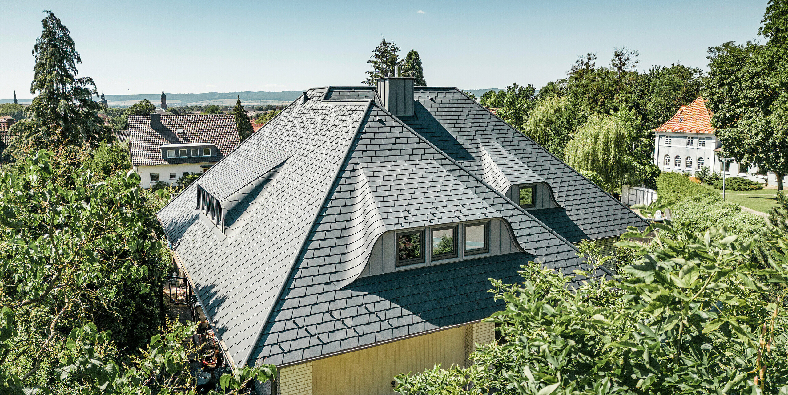 Blick auf das Dach eines traditionellen Einfamilienhauses in Einbeck, Deutschland. Die einzigartige Dachlandschaft wurde mit robusten PREFA Dachschindeln in P.10 Anthrazit eingedeckt. Das Dach zeichnet sich durch seine anmutigen Hechtgauben und die präzise Verarbeitung der Schindeln aus, die sich harmonisch in die ländliche Umgebung und das grüne Landschaftsbild einfügen. Die Kombination aus zeitgenössischem Dachmaterial und klassischer Architektur verbindet Tradition mit Innovation und Nachhaltigkeit.