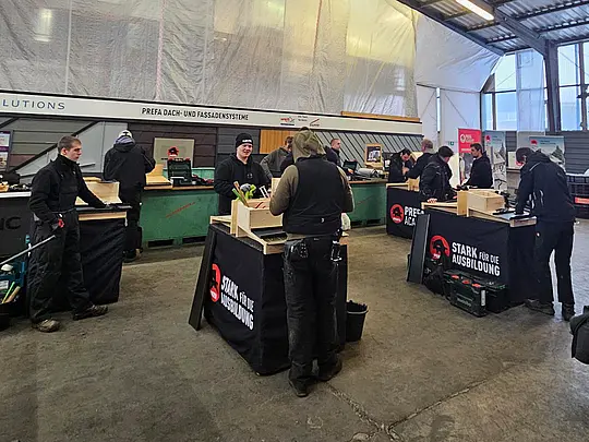 PREFA Academy Cube in einer Werkhalle: Mehrere Teilnehmer arbeiten gleichzeitig an Trainingsstationen und montieren PREFA Produkte an eine Holz-UK. Werkzeuge, Materialboxen und PREFA Branding sind im Hintergrund sichtbar.