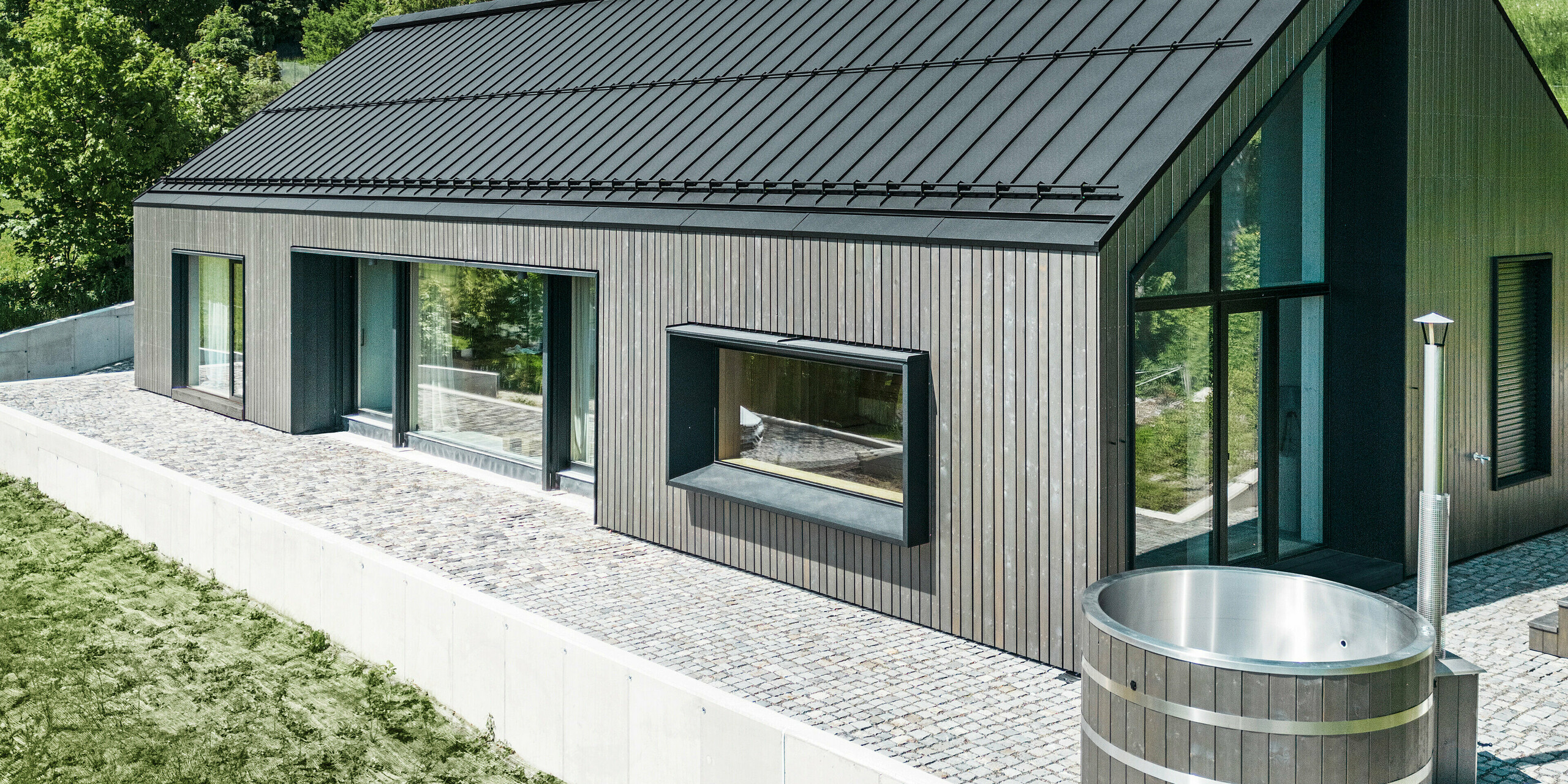 Ein modernes Holzmassivhaus in Kuncice (CZ) wurde mit großen Fensterflächen ausgestattet. Das dunkle Holzmassivhaus verfügt über ein Dachsystem aus PREFALZ in der Farbe P. 10 Schwarz. Im hinteren Außenbereich steht ein Whirlpool mit einer Holzverkleidung.