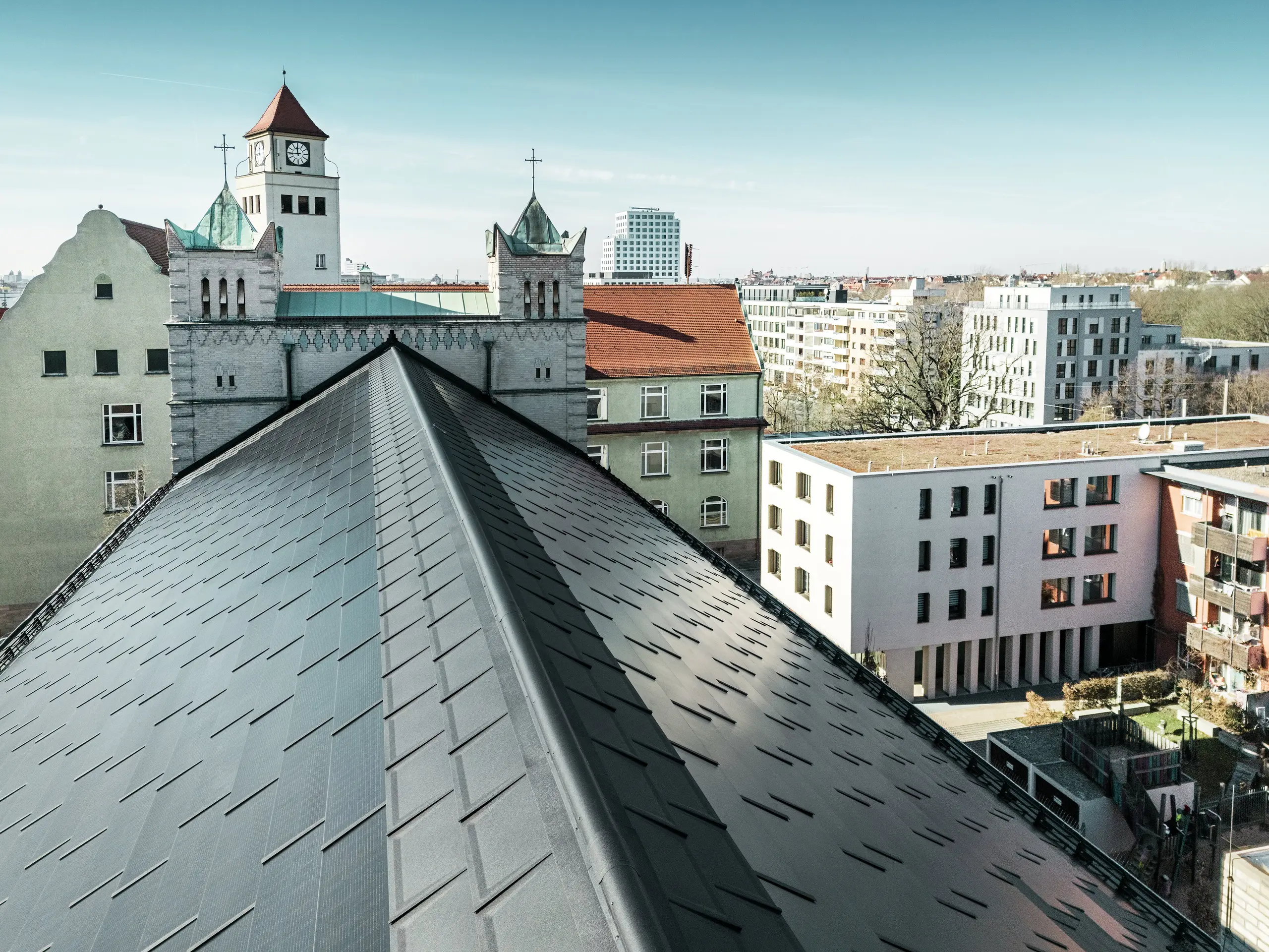 Das Bild zeigt die präzise Deckung des Aluminiumdachs der St. Karl Borromäus Kirche in Nürnberg. Das Kirchendach wurde mit PREFA Solardachplatten in elegantem Schwarz ausgestattet. Der Blick auf den markanten First des Satteldachs unterstreicht die gelungene Integration der modernen Photovoltaik-Indach-Lösung in die historische Architektur. Die Solardachplatten fügen sich harmonisch auf dem denkmalgeschützten Gebäude ein und versorgen die Kirche und etliche benachbarte Gebäude mit effizienter und nachhaltiger Energie.