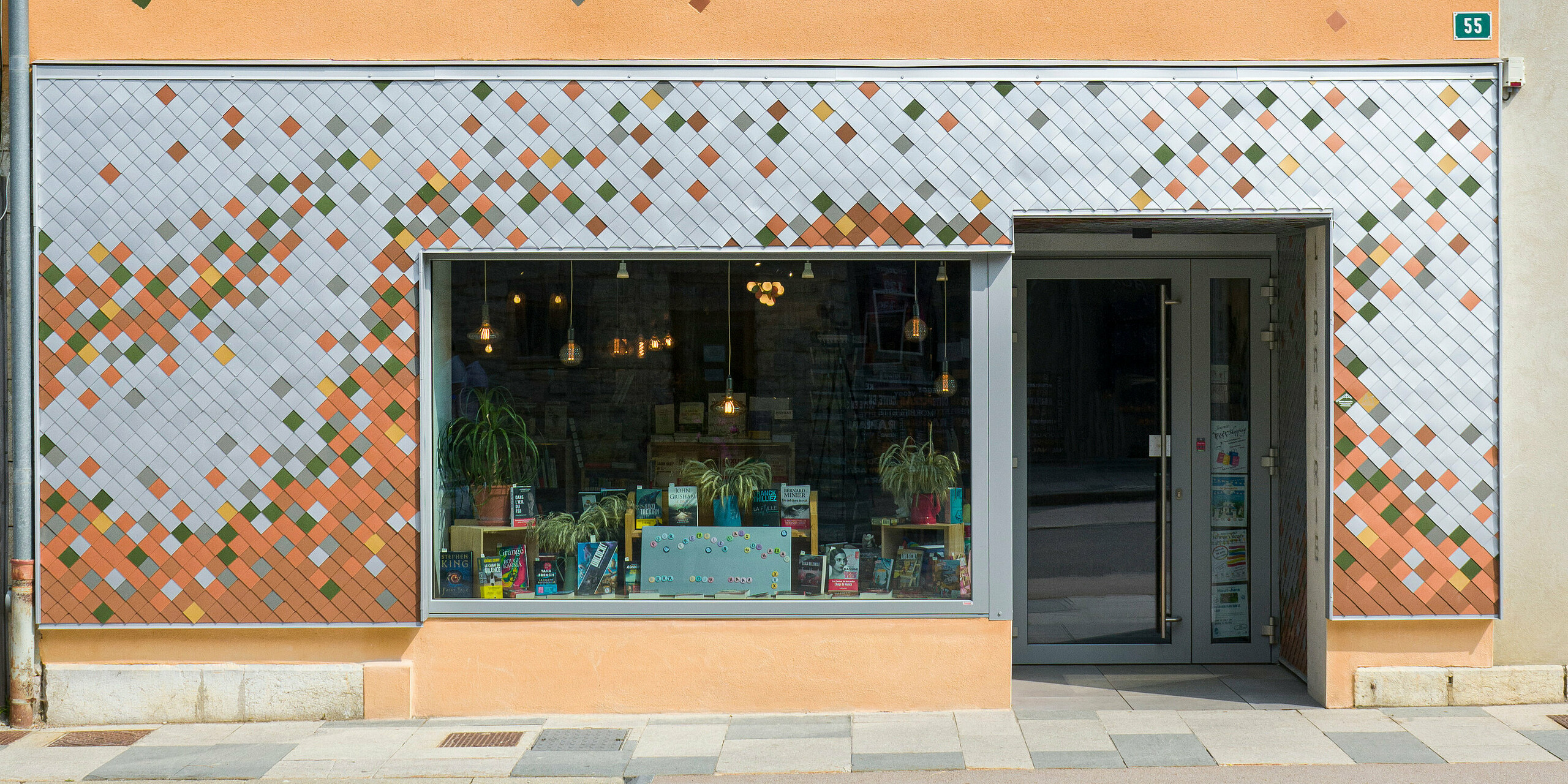 Frontansicht der bunten Fassade einer Buchhandlung in der kleinen französischen Gemeinde Les Rousses. Das Gebäude kombiniert ein leuchtend orangefarbenes Obergeschoss mit dekorativen Elementen und ein unteres Geschoss mit einer mosaikartigen Fassade aus Aluminium in verschiedenen Farben. Ein großes Schaufenster im Erdgeschoss präsentiert Bücher und Pflanzen neben der Eingangstür. Eine schmale Dachrinne verläuft an der Seite des Gebäudes, und die Nummer 55 ist neben der Tür auf einem grünen Schild zu sehen.