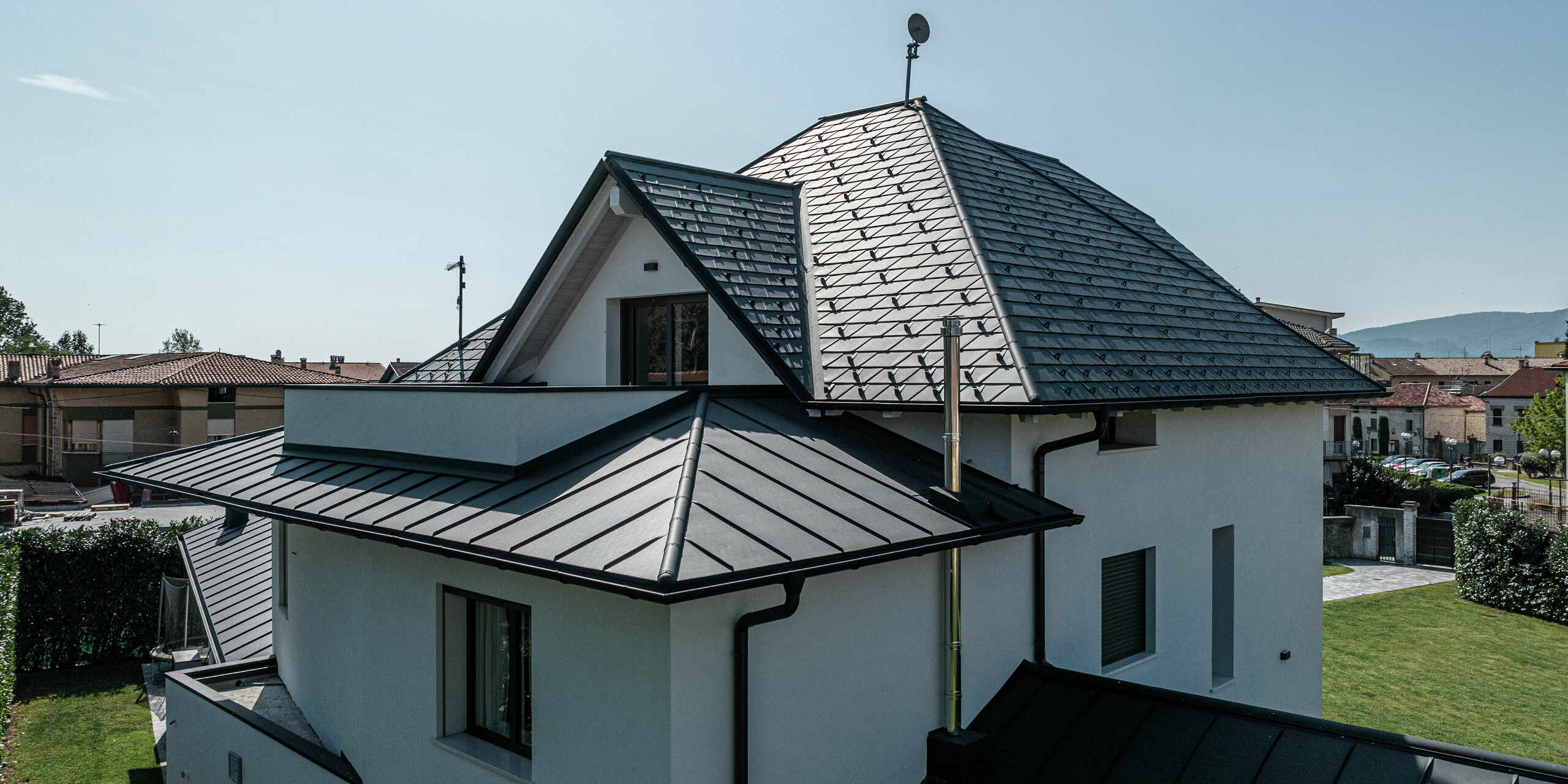 Detailansicht der Dachrückseite eines modernen Einfamilienhauses in Marano Vicentino, Italien. Das Hauptdach ist mit PREFA Dachschindeln in P.10 Anthrazit eingedeckt, während die beiden Anbauten mit einer eleganten Doppelstehfalzdeckung aus Aluminium versehen sind. Die Kombination hochwertiger Aluminiumprodukte sorgt für eine langlebige, witterungsbeständige und ästhetische Dachlösung.