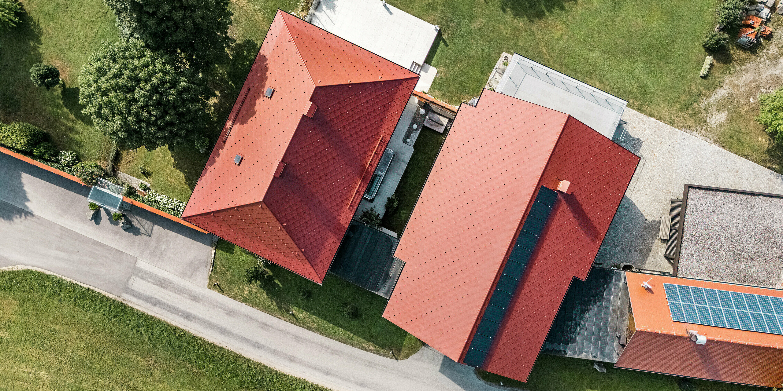 Die Vogelperspektive zeigt die beiden sanierten Einfamilienhäuser in Aschach an der Steyr, deren Dächer mit PREFA Dachrauten 44 x 44 in der Farbe P.10 Oxydrot gedeckt wurden. Die rote Farbe der hochwertigen Blechdächer setzt einen markanten Akzent und harmoniert perfekt mit der Umgebung. Die präzise Anordnung der Dachrauten und die einheitliche Gestaltung betonen die moderne und gleichzeitig traditionelle Architektur. 