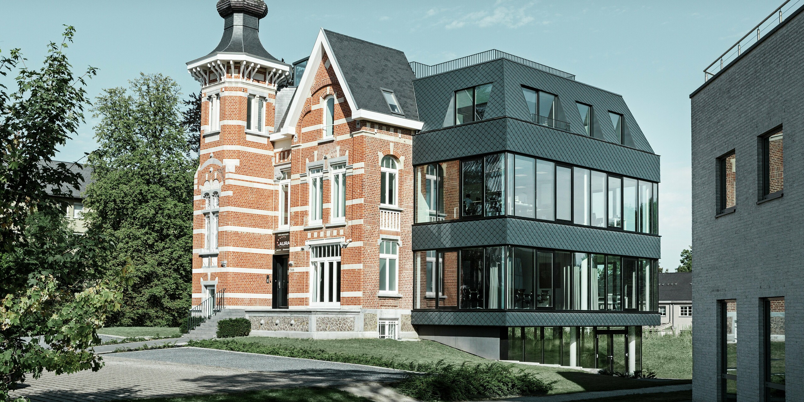 Aus der seitlichen Perspektive zeigt sich die eindrucksvolle Symbiose von historischer Backsteinvilla mit Turm und modernem Zubau aus Glas und PREFA Wandrauten in Anthrazit. Die klar strukturierte Aluminiumfassade der Villa Kasteeltje Heizel setzt mit ihrer markanten Rautenoptik architektonische Akzente, während große Fensterflächen Transparenz und Leichtigkeit schaffen. Tradition trifft Innovation in perfekter Harmonie.