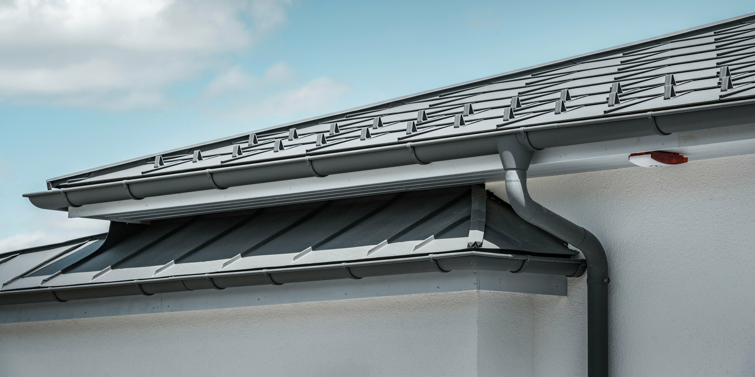 Die Detailaufnahme eines Einfamilienhauses in Ilmenau, Thüringen, zeigt das effiziente PREFA Dachentwässerungssystem in P.10 Dunkelgrau. Das Bild fokussiert auf die nahtlose Integration der Hängerinne und des Ablaufrohrs, die eine optimale Ableitung von Regenwasser gewährleisten. Die dunkelgraue Stehfalzdeckung aus PREFALZ und die Dachplatten R.16, ebenfalls in Dunkelgrau, runden das hochwertige Gesamtbild des Daches ab und spiegeln die Kombination aus Funktionalität und anspruchsvollem Design wider.