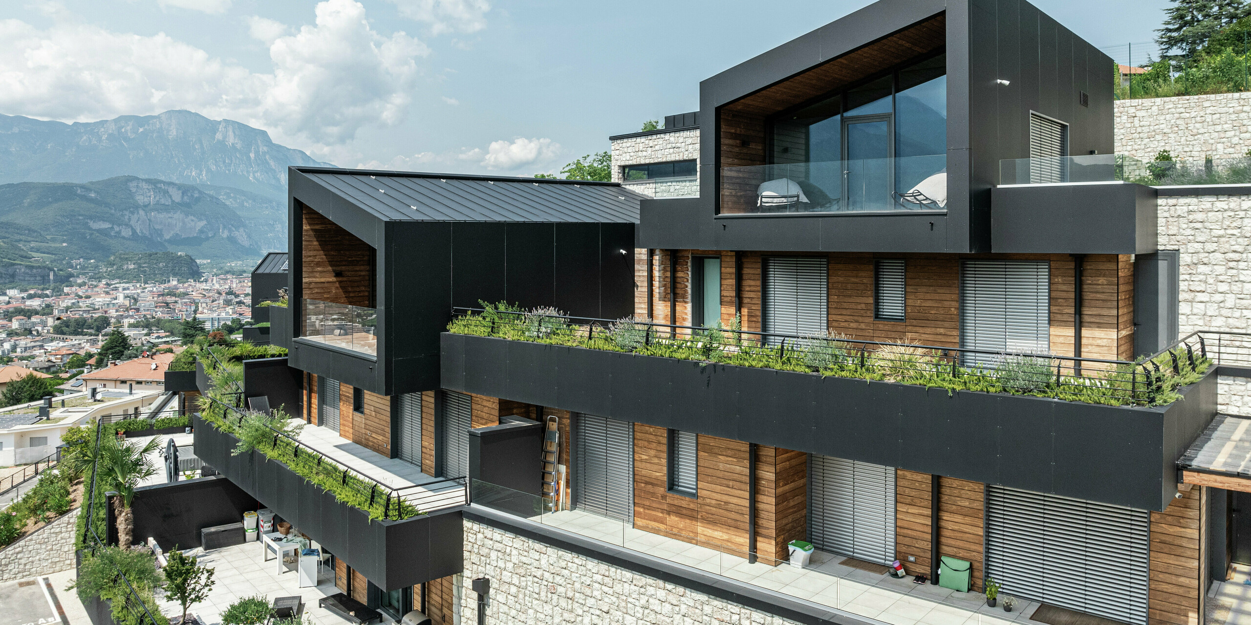 Moderne Wohnarchitektur mit Charakter: Die elegante Fassade aus PREFA Aluminium Verbundplatten in P.10 Schwarz harmoniert perfekt mit den warmen Holzelementen und dem Natursteinsockel. Das PREFALZ Dach in P.10 Schwarz bietet nicht nur eine ästhetische Einheit, sondern auch höchste Witterungsbeständigkeit. Großzügige Glasflächen und begrünte Terrassen schaffen eine perfekte Verbindung zwischen Innen- und Außenraum, während die Hanglage einen beeindruckenden Blick über Trient ermöglicht. Langlebig, pflegeleicht und stilvoll - ein Wohnkonzept mit Zukunft.