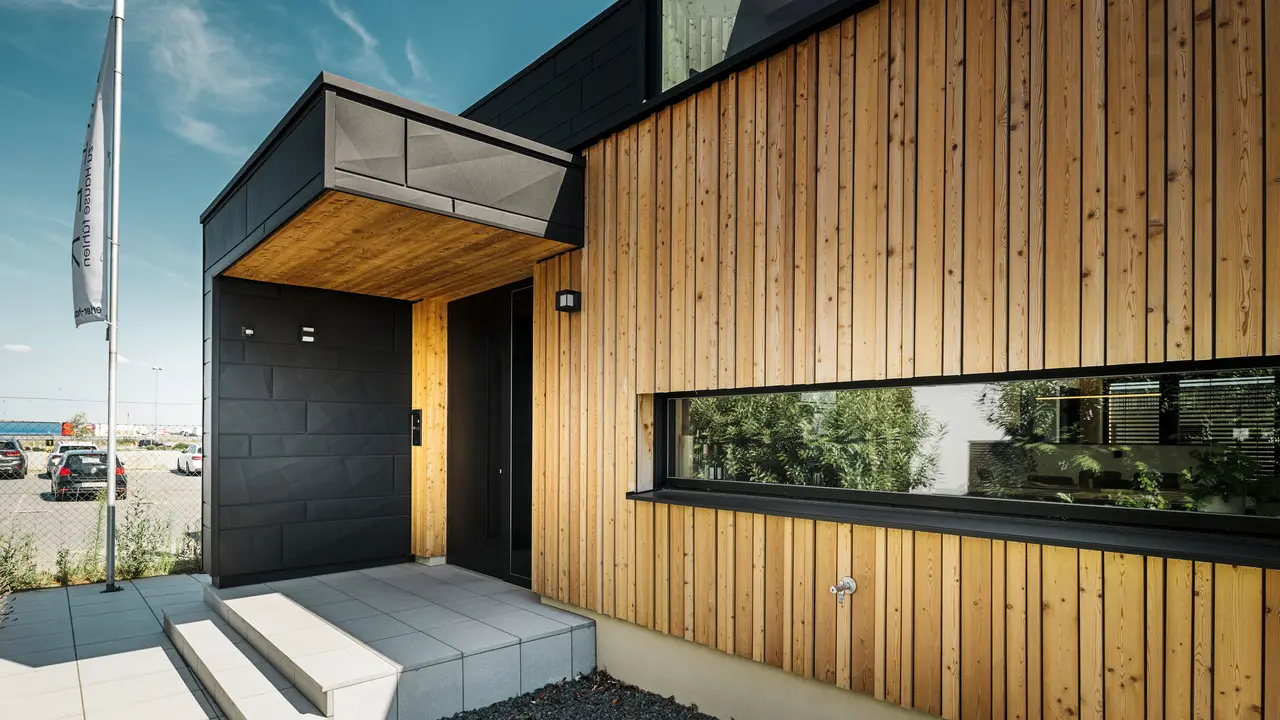Seitlicher Eingang des Musterhauses Autark im Fertighauszentrum Blaue Lagune in Vösendorf, mit einer modernen Fassade aus natürlichem Holz und schwarzen PREFA Siding.X Paneelen. Das Bild zeigt die edle Kombination aus Holzverkleidung und Aluminium in der hochwertigen Beschichtung P.10 Schwarz.