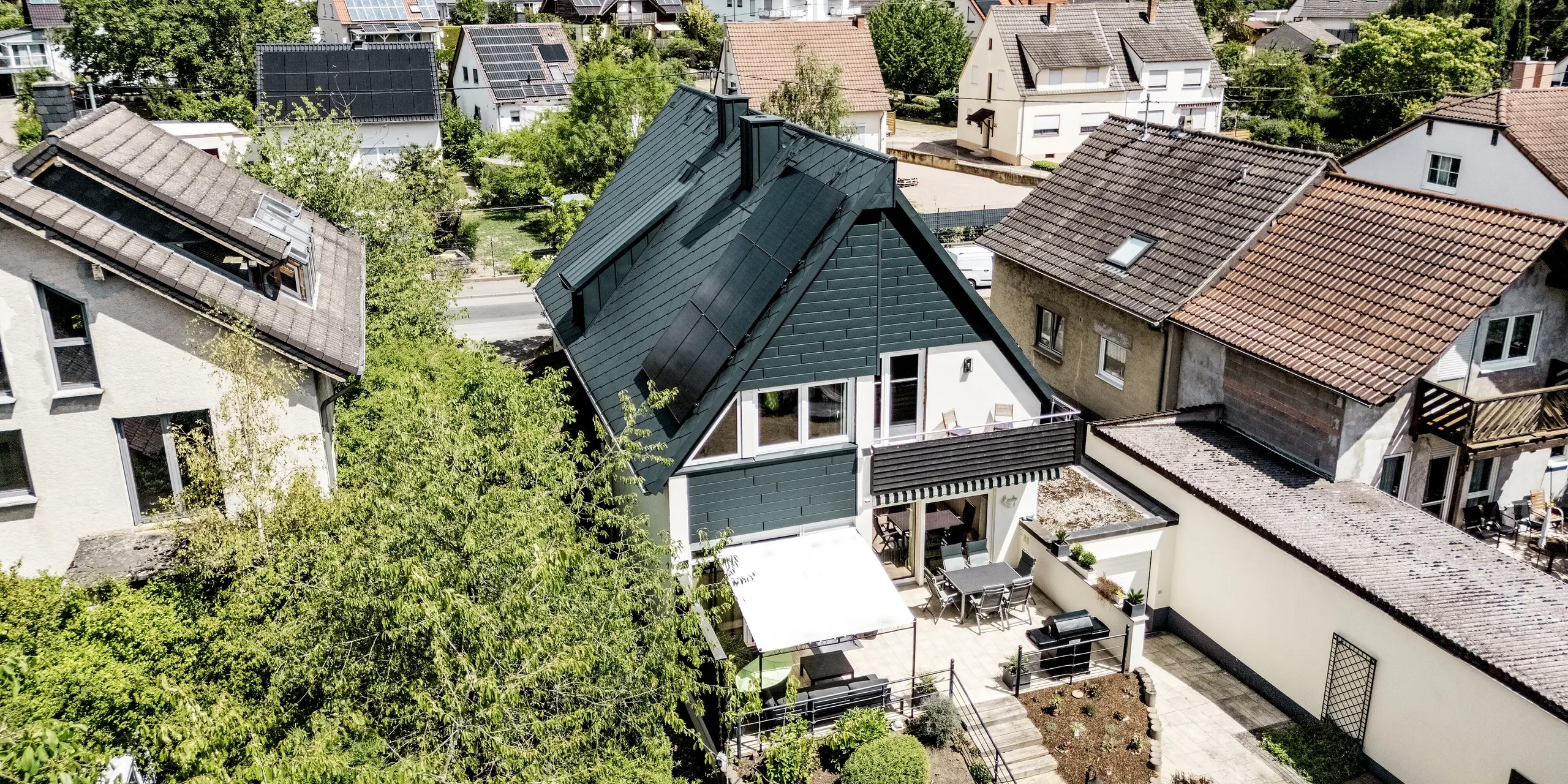 Luftaufnahme eines sanierten Einfamilienhauses in Hargesheim, das mit seinem anthrazitfarbenen PREFA Aluminiumdach aus Dachrauten 44 × 44 hervorsticht. Die mit Siding.X verkleidete Giebelfassade in P.10 Anthrazit schafft einen modernen Kontrast zur hellen Putzfassade. Langlebige, leichte und witterungsbeständige Aluminiumprodukte verleihen dem Haus inmitten der grünen Wohnsiedlung einen unverwechselbaren Charakter.