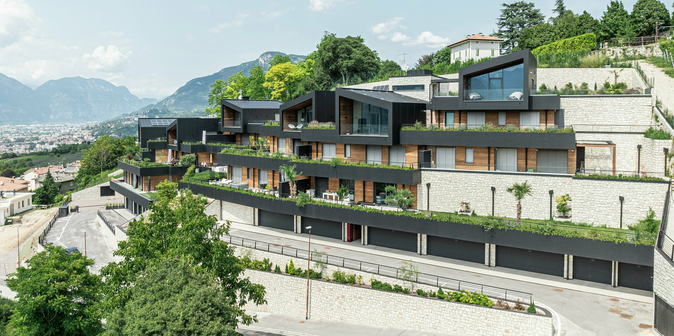 Die Wohnanlage fügt sich perfekt in die Hanglage oberhalb von Trient ein und bietet einen beeindruckenden Panoramablick auf die Stadt und die umliegende Berglandschaft. Die Kombination aus Naturstein, Holz und der hochwertigen PREFA Aluminiumfassade in P.10 Schwarz sorgt für ein modernes, elegantes Erscheinungsbild mit höchster Witterungsbeständigkeit. Das PREFALZ Dach in P.10 Schwarz garantiert Langlebigkeit und Schutz. Großzügige Terrassen mit gepflegter Bepflanzung unterstreichen das nachhaltige Wohnkonzept und schaffen ein harmonisches Zusammenspiel von Architektur und Natur.