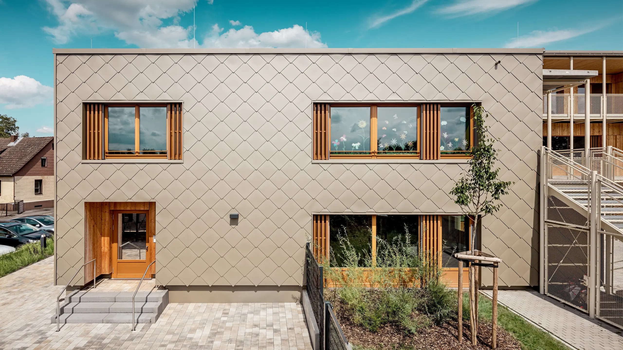 Die Rückseite der modernen Kindertagesstätte in Alzenau wird aus einer frontalen Perspektive gezeigt und beeindruckt mit klaren Linien und einer markanten Fassade aus bronzefarbenen PREFA Wandrauten 44 × 44. Die geometrische Fassadenstruktur erzeugt ein harmonisches Schuppenmuster, das durch die ruhige, horizontale Ausrichtung der Fenster akzentuiert wird. Großzügige Holzrahmen und vertikale Lamellen setzen natürliche Akzente und sorgen für eine warme, einladende Atmosphäre. Die durchdachte Kombination aus langlebigem Aluminium und regionalem Holz steht für nachhaltige Architektur mit hoher gestalterischer Qualität. Das Bild betont die präzise Ausführung, die hochwertige Materialwahl und die gelungene Integration des Gebäudes in das städtische Umfeld.