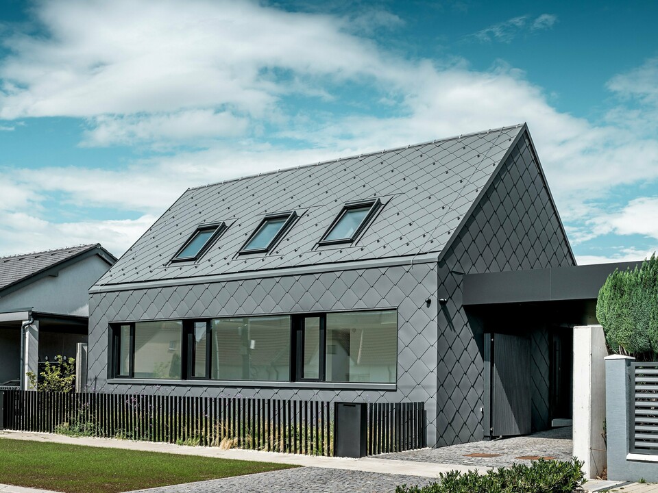 Modernes Einfamilienhaus in Trnava‑Modranka mit Aluminiumhülle: Die gesamte Gebäudehülle dieses schlichten Gebäudes wurde mit PREFA Dach- und Wandrauten 44 × 44 in P.10 Dunkelgrau gestaltet. Die markante Rautenstruktur zieht sich nahtlos über Dach und Fassade und verleiht dem Haus eine starke, monolithische Wirkung. Große Fensteröffnungen setzen klare architektonische Akzente, während das witterungsbeständige PREFA Aluminium langlebigen Schutz und eine edle, matte Optik bietet. Der helle Himmel verstärkt den Kontrast zur dunklen Gebäudehülle und betont das moderne Design.