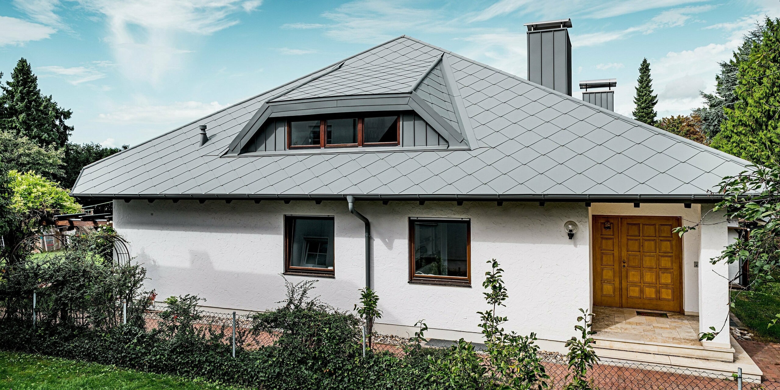 Das Bild zeigt ein modern saniertes Einfamilienhaus mit einem markanten Walmdach, das vollständig mit PREFA Dachrauten 44 × 44 in P.10 Hellgrau eingedeckt ist. Die Stucco-Struktur verleiht der Dachfläche eine elegante, lebendige Optik. Die sauber integrierte Trapezgaube und die hellgraue Kaminverkleidung schaffen ein harmonisches Gesamtbild. Die farblich abgestimmte PREFA Dachentwässerung unterstreicht die hochwertige Ausführung. Die langlebigen, leichten Aluminiumprodukte bieten höchste Stabilität und machen das Dach besonders widerstandsfähig gegen Witterungseinflüsse.