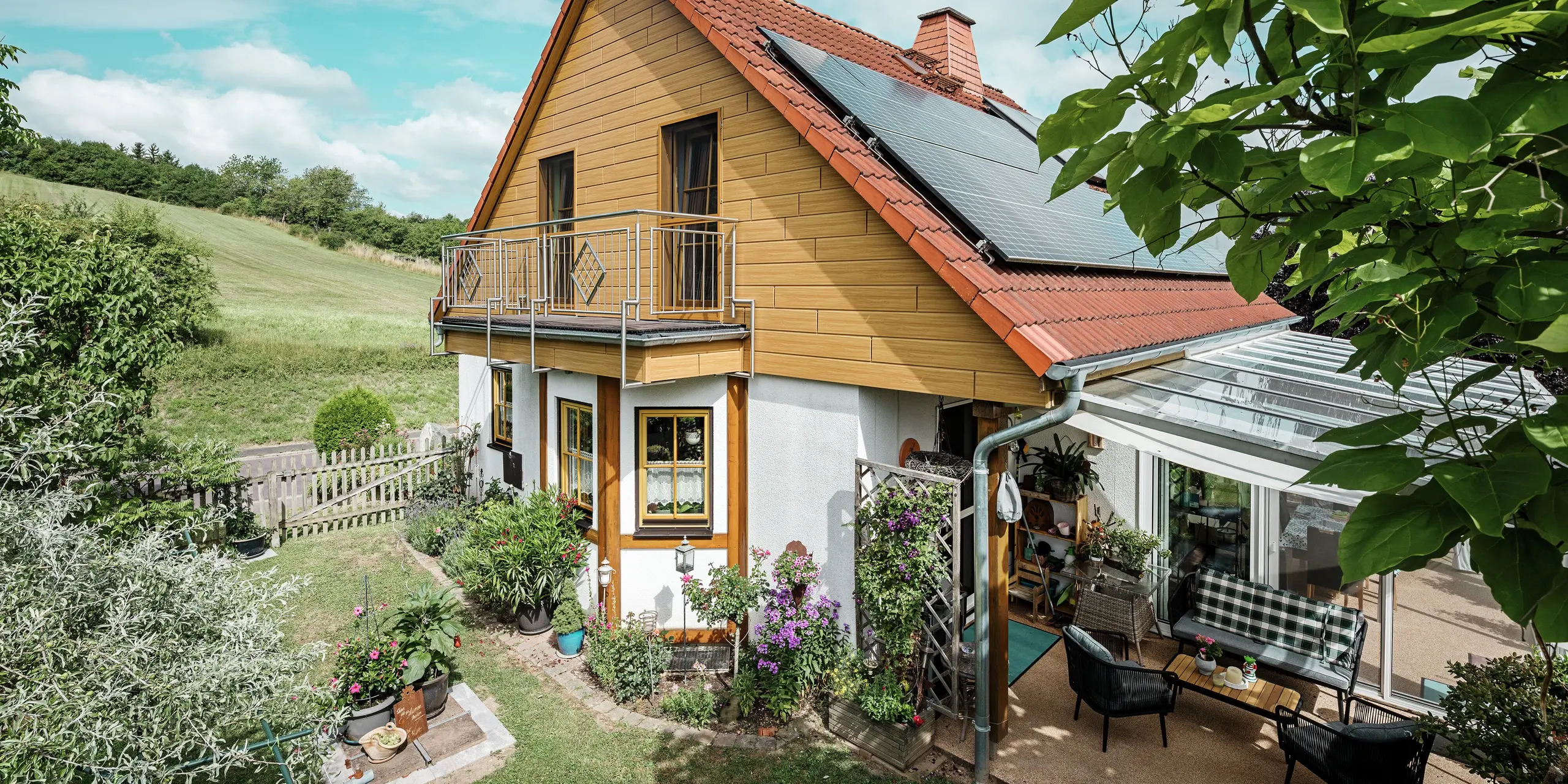 Dieses Bild zeigt ein charmantes Einfamilienhaus in Blieskastel, dessen frisch sanierter Giebel mit PREFA Sidings in Eiche Natur sofort ins Auge fällt. Die warme Holzoptik aus langlebigem Aluminium harmoniert beeindruckend mit dem roten Ziegeldach und dem großzügigen Wintergarten. Umgeben von üppigem Grün und liebevoll angelegten Blumenbeeten entsteht eine idyllische, lebendige Szene. Die robuste Fassadensanierung mit PREFA verleiht dem Haus nicht nur einen natürlichen Look, sondern überzeugt auch durch Witterungsbeständigkeit, Farbtreue und nahezu wartungsfreie Langlebigkeit.
