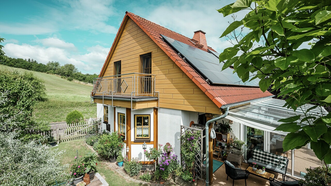 Dieses Bild zeigt ein charmantes Einfamilienhaus in Blieskastel, dessen frisch sanierter Giebel mit PREFA Sidings in Eiche Natur sofort ins Auge fällt. Die warme Holzoptik aus langlebigem Aluminium harmoniert beeindruckend mit dem roten Ziegeldach und dem großzügigen Wintergarten. Umgeben von üppigem Grün und liebevoll angelegten Blumenbeeten entsteht eine idyllische, lebendige Szene. Die robuste Fassadensanierung mit PREFA verleiht dem Haus nicht nur einen natürlichen Look, sondern überzeugt auch durch Witterungsbeständigkeit, Farbtreue und nahezu wartungsfreie Langlebigkeit.
