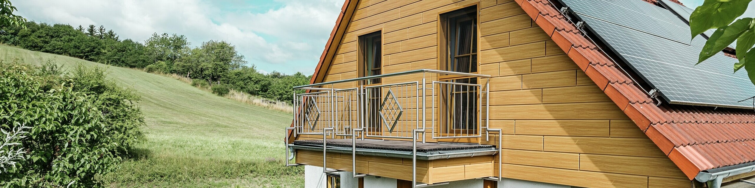 Dieses Bild zeigt ein charmantes Einfamilienhaus in Blieskastel, dessen frisch sanierter Giebel mit PREFA Sidings in Eiche Natur sofort ins Auge fällt. Die warme Holzoptik aus langlebigem Aluminium harmoniert beeindruckend mit dem roten Ziegeldach und dem großzügigen Wintergarten. Umgeben von üppigem Grün und liebevoll angelegten Blumenbeeten entsteht eine idyllische, lebendige Szene. Die robuste Fassadensanierung mit PREFA verleiht dem Haus nicht nur einen natürlichen Look, sondern überzeugt auch durch Witterungsbeständigkeit, Farbtreue und nahezu wartungsfreie Langlebigkeit.