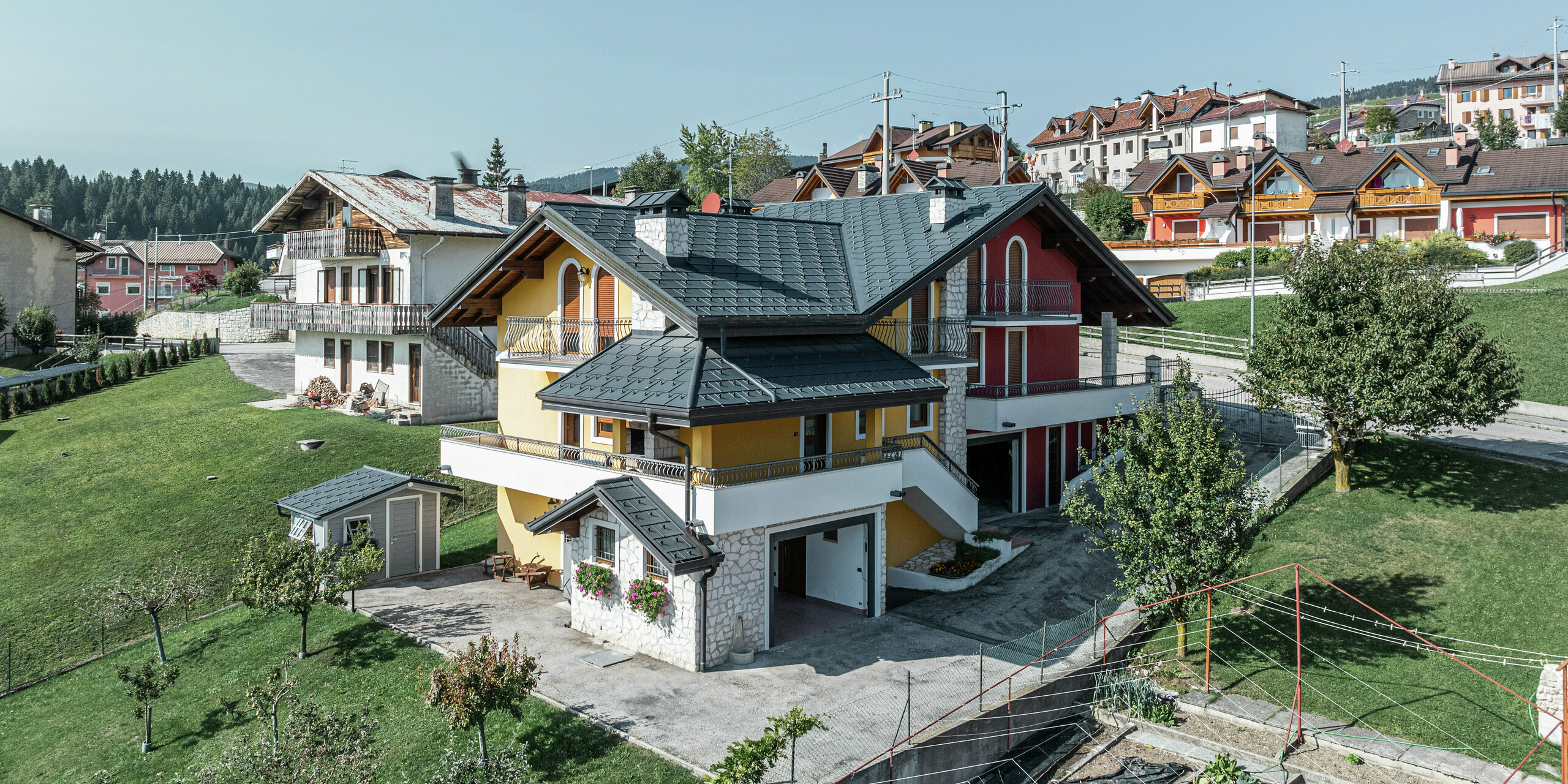 Einfamilienhaus in Gallio, Italien, im traditionellen Alpenstil mit modernem PREFA Dach in P.10 Dunkelgrau. PREFA Dachplatten R.16 bieten optimalen Schutz vor Schnee, Regen und Wind. Die einzigartige Kombination aus gelb-roter Fassade, Natursteinelementen und Holzakzenten verleiht dem Haus eine einladende Optik. Die PREFA Dachentwässerung sorgt für eine zuverlässige Ableitung der Niederschläge. Eingebettet in eine grüne Hügellandschaft verbindet dieses Haus einen alpinen Charme mit langlebiger, wartungsarmer Dachtechnik.