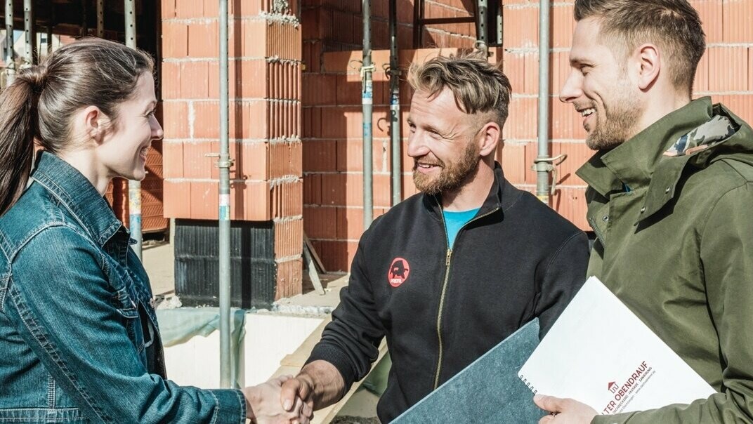 Ein junges Pärchen trifft sich auf ihrer Hausbau-Baustelle mit dem Handwerker in der Nähe. Sie haben das Angebot besprochen und angenommen. Der Spengler hält eine PREFA Dachplatte in der Hand.