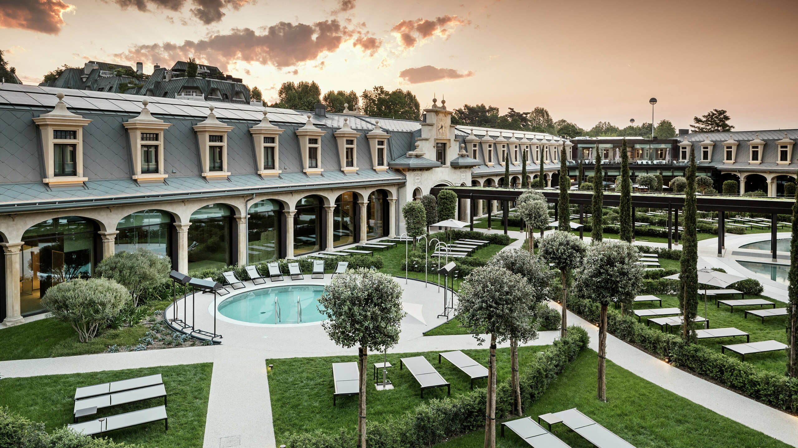 Abendstimmung im Thermalpark „De Montel – Terme Milano“ mit eindrucksvollem Blick auf die denkmalgeschützte Fassadenarchitektur im Jugendstil. Die Dacheindeckung mit PREFA Dachrauten 44 × 44 in P.10 Hellgrau verbindet Nachhaltigkeit mit Ästhetik. Im Vordergrund laden Liegewiesen, gepflegte Wege, Olivenbäume und ein rundes Thermalbecken zum Verweilen ein.