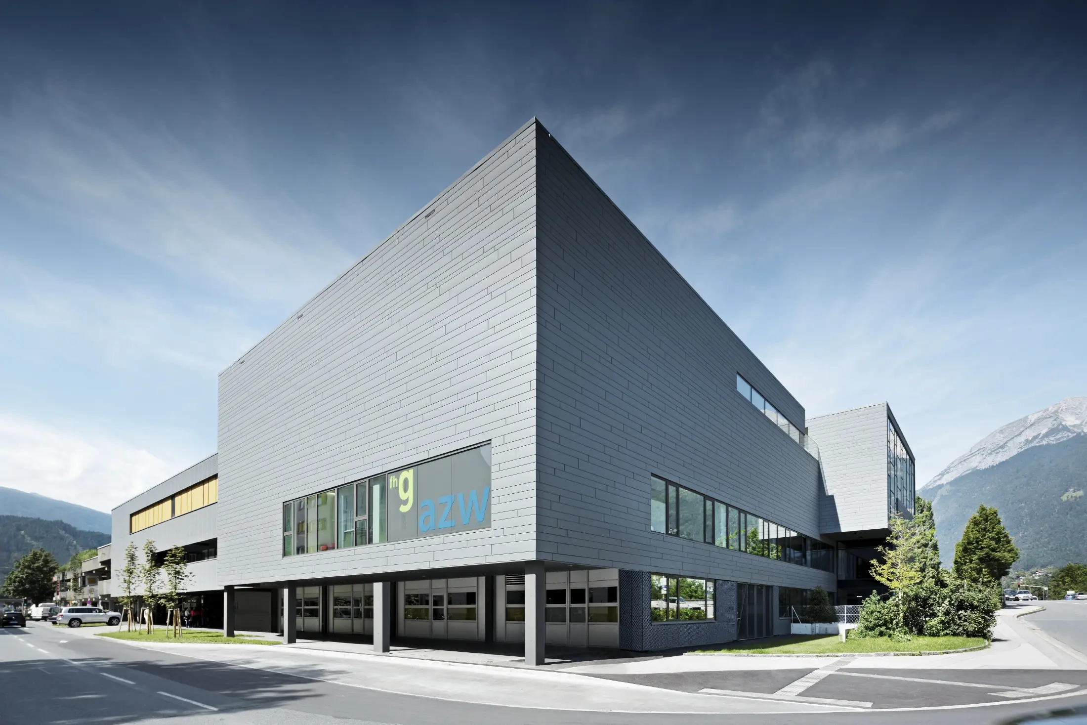 Modernes Ausbildungszentrum mit klarer, kubischer Architektur und hellgrauer Aluminiumfassade. Das Gebäude hat große Fensterflächen und überhängende Baukörper. Auf einer Glasfront ist die Beschriftung „fhg azw“ zu sehen. Im Hintergrund erheben sich Berge unter einem leicht bewölkten Himmel, während im Vordergrund eine gepflegte Straße mit Grünflächen verläuft.
