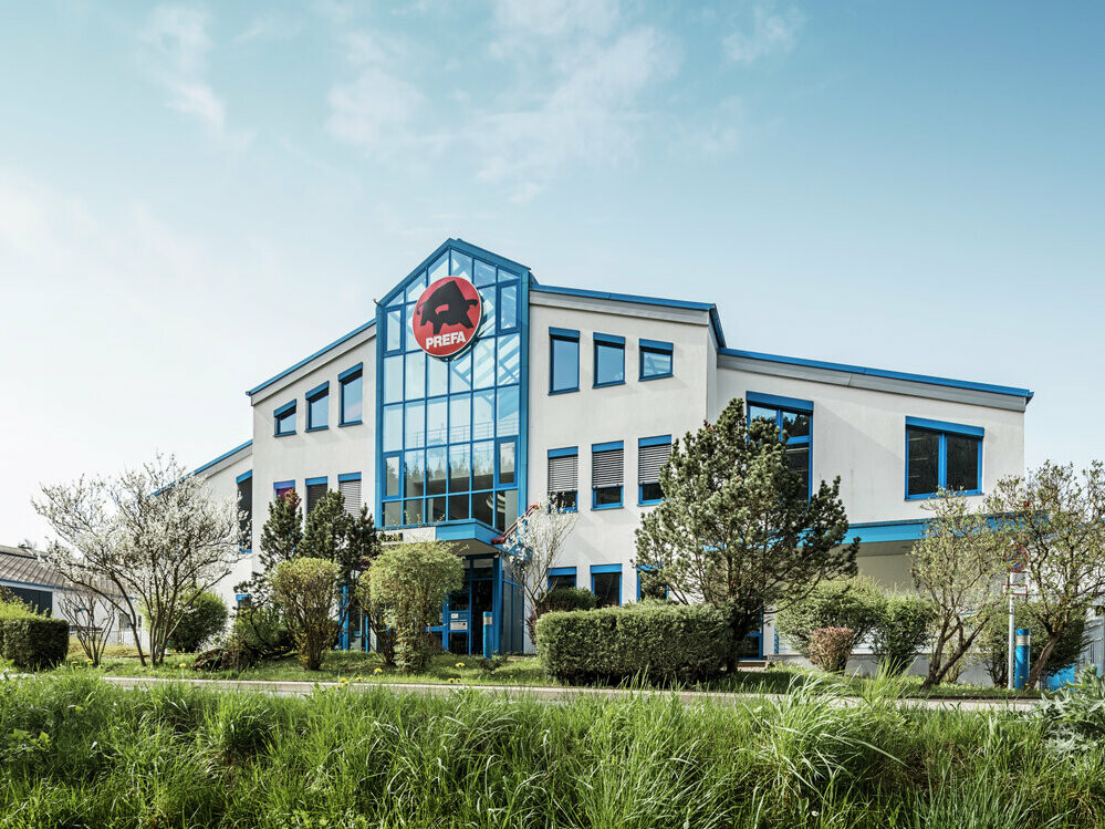 Das PREFA Firmengebäude in Wasungen, Deutschland, ist ein modernes, helles Gebäude mit blauen Fensterrahmen und einer markanten Glasfront. Über dem Eingang befindet sich das rote PREFA-Logo mit dem schwarzen Stier. Das Gebäude ist von gepflegten Grünflächen und Bäumen umgeben.