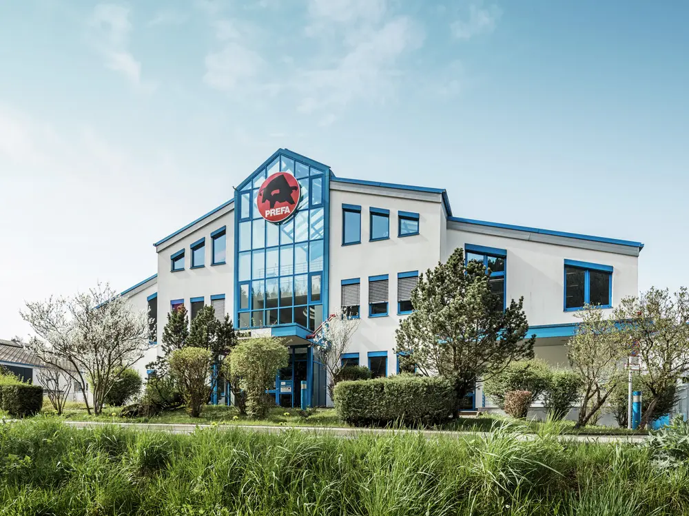 Das PREFA Firmengebäude in Wasungen, Deutschland, ist ein modernes, helles Gebäude mit blauen Fensterrahmen und einer markanten Glasfront. Über dem Eingang befindet sich das rote PREFA-Logo mit dem schwarzen Stier. Das Gebäude ist von gepflegten Grünflächen und Bäumen umgeben.