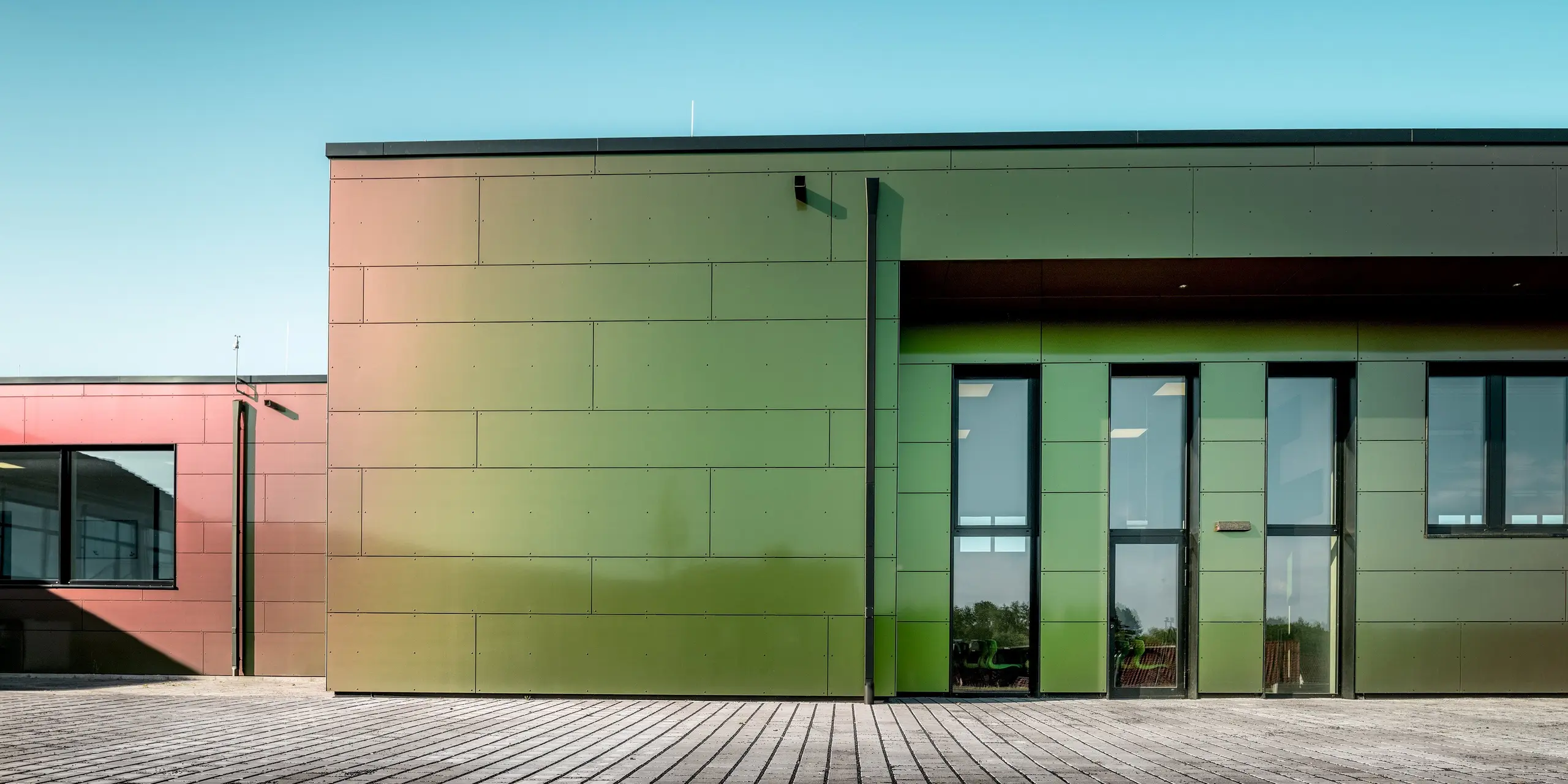 Faszinierendes Farbspiel an der Johannwolfschule Außenstelle Angerstein: Großformatige PREFA Aluminium Verbundplatten in der Sonderfarbe Spring Lake lassen die moderne Fassade je nach Lichteinfall in Grün-, Bronze- und Rottönen schimmern. Die langlebigen, witterungsbeständigen Aluminiumplatten betonen die geradlinige Architektur, während das PREFA Quadratrohr die elegante Dachentwässerung übernimmt.
