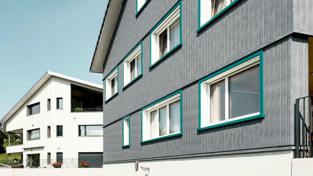 Ansicht zweier benachbarter Mehrfamilienhäuser in Weissbad, Schweiz, wobei das vordere Gebäude mit seiner PREFA Siding Fassade in P.10 Steingrau einen starken visuellen Eindruck vermittelt. Die vertikal montierten Sidings schaffen eine elegante und schützende Hülle, während die türkisen Fensterrahmen farbliche Akzente setzen. Das Bild fängt das Zusammenspiel der modernen Materialien mit der traditionellen Architektur ein und demonstriert die vielseitige Einsetzbarkeit von PREFA Aluminiumprodukten.