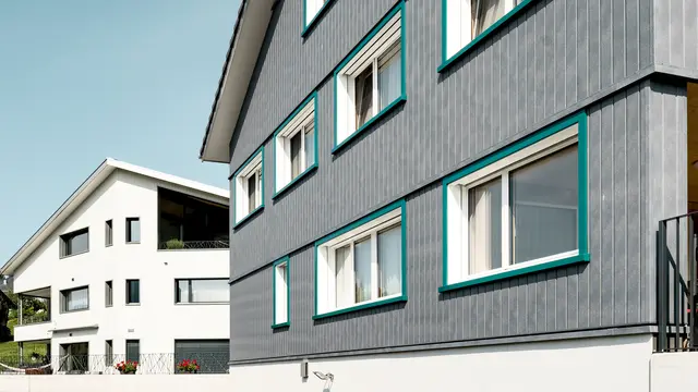 Ansicht zweier benachbarter Mehrfamilienhäuser in Weissbad, Schweiz, wobei das vordere Gebäude mit seiner PREFA Siding Fassade in P.10 Steingrau einen starken visuellen Eindruck vermittelt. Die vertikal montierten Sidings schaffen eine elegante und schützende Hülle, während die türkisen Fensterrahmen farbliche Akzente setzen. Das Bild fängt das Zusammenspiel der modernen Materialien mit der traditionellen Architektur ein und demonstriert die vielseitige Einsetzbarkeit von PREFA Aluminiumprodukten.