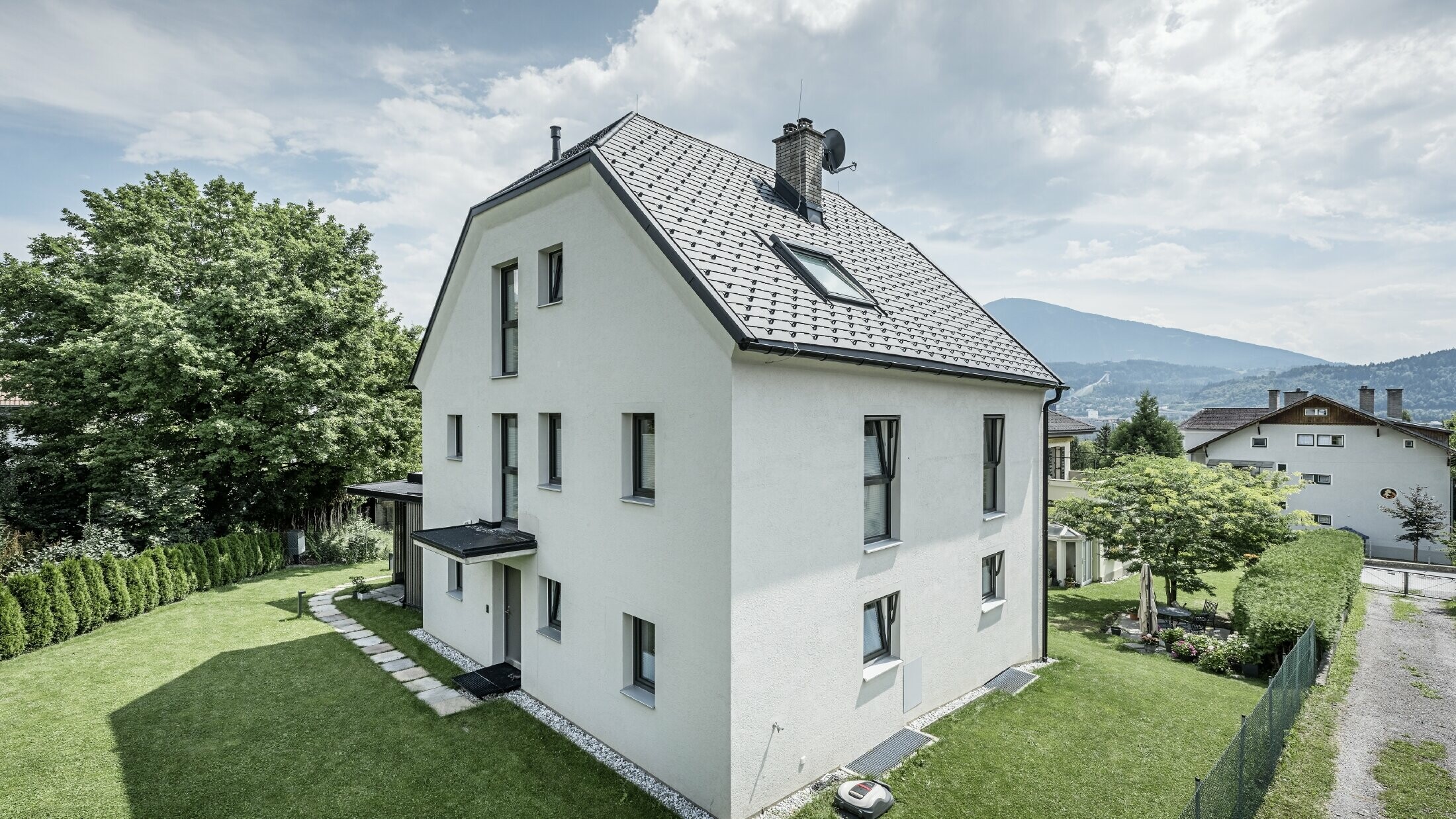 Modernes dreigeschossiges Einfamilienhaus mit heller Putzfassade und dunkelgrauem Satteldach aus Aluminium von PREFA. Das Gebäude verfügt über mehrere schmale Fenster und eine Dachentwässerung in Anthrazit. Es steht in einem gepflegten Garten mit Rasenfläche, Bäumen und Hecken, im Hintergrund sind weitere Wohnhäuser und eine bergige Landschaft zu sehen.