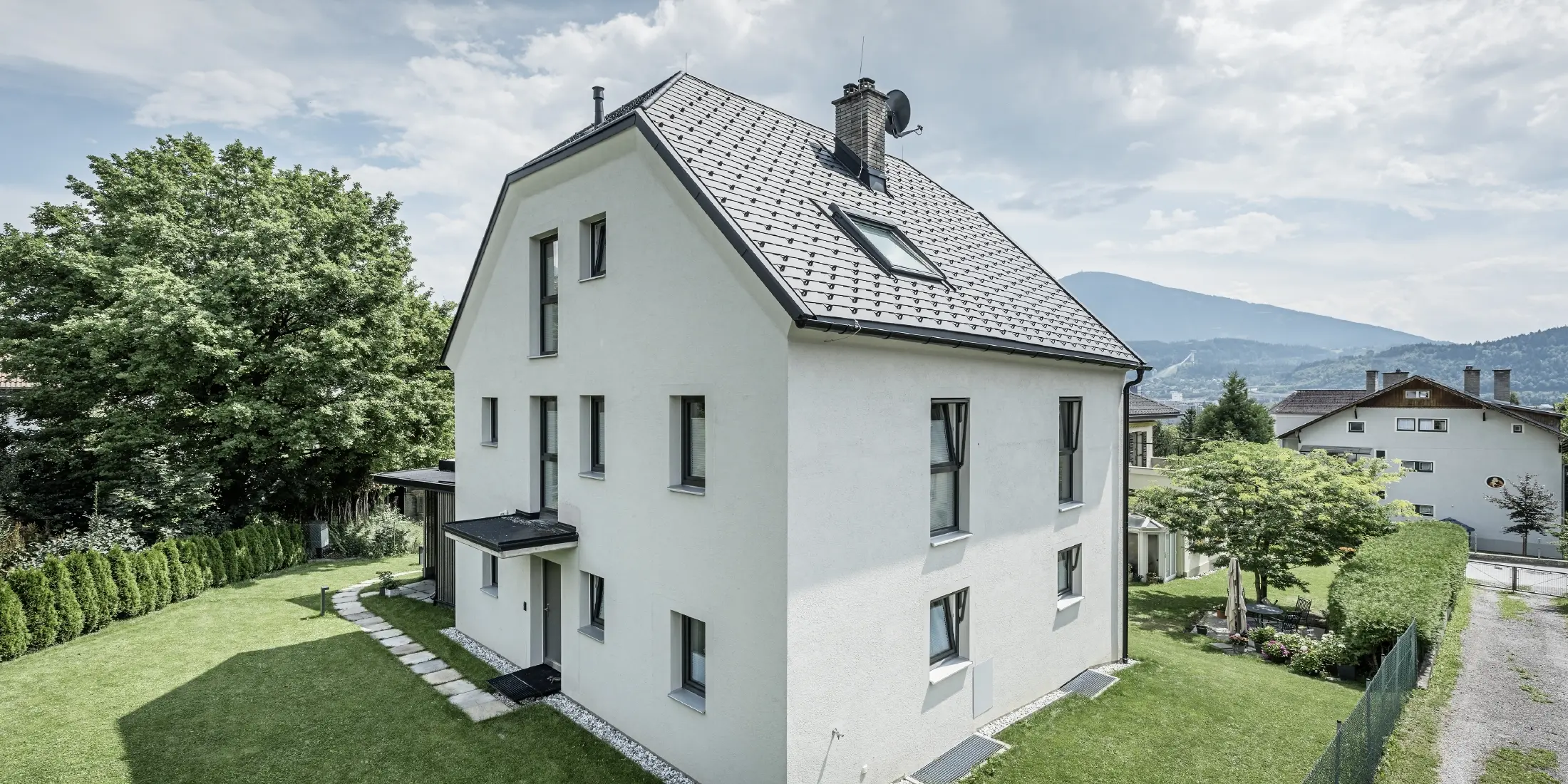 Modernes dreigeschossiges Einfamilienhaus mit heller Putzfassade und dunkelgrauem Satteldach aus Aluminium von PREFA. Das Gebäude verfügt über mehrere schmale Fenster und eine Dachentwässerung in Anthrazit. Es steht in einem gepflegten Garten mit Rasenfläche, Bäumen und Hecken, im Hintergrund sind weitere Wohnhäuser und eine bergige Landschaft zu sehen.