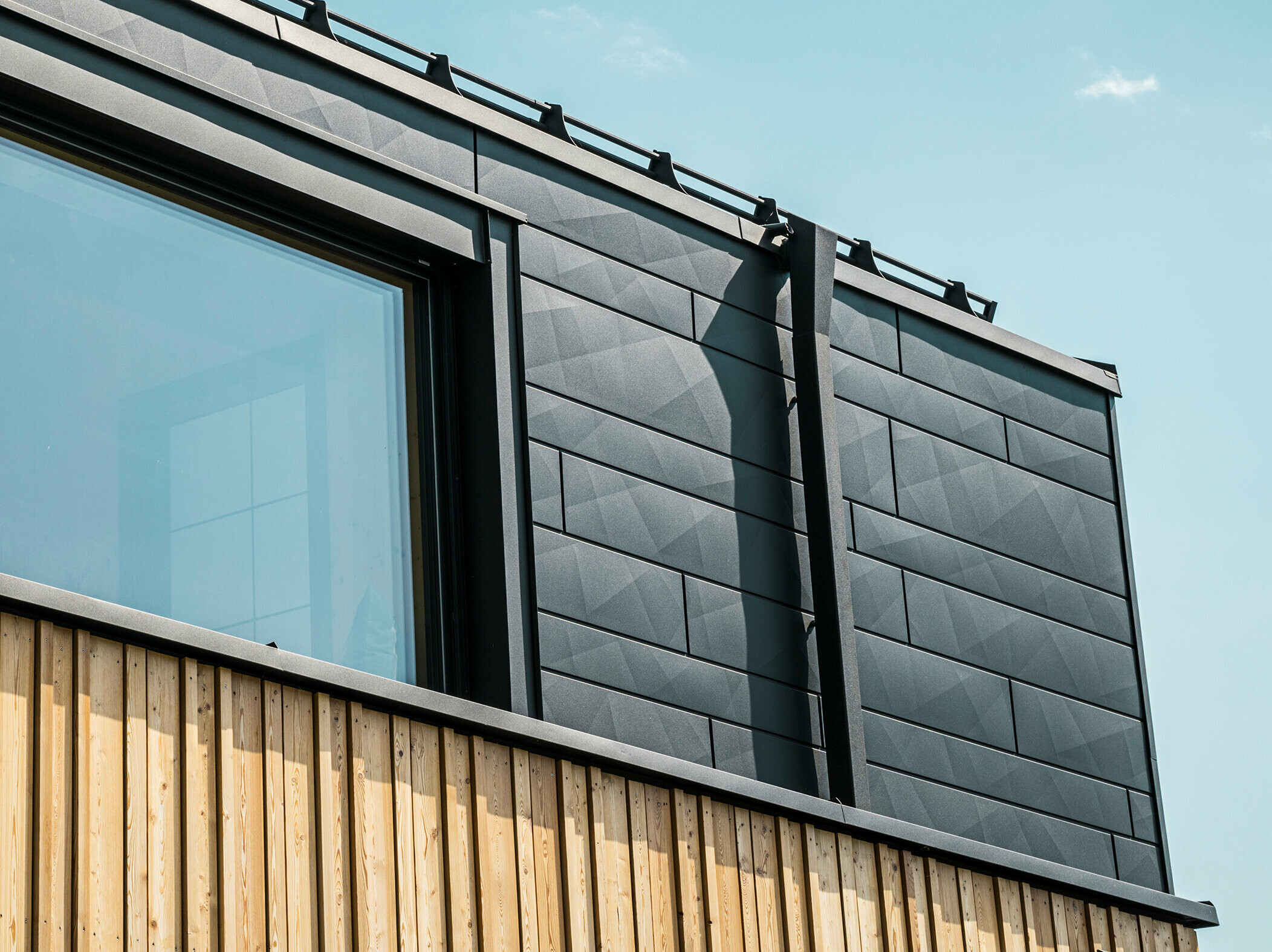 Nahaufnahme des Obergeschosses des Musterhauses Autark im Fertighauszentrum Blaue Lagune, Vösendorf (AT). Die Fassade besteht aus PREFA Siding.X Aluminiumpaneelen in P.10 Schwarz, die durch ihre spezielle Optik mit unterschiedlichen Kantungen auffallen. Darunter befindet sich eine stilvolle Holzverkleidung, die einen harmonischen Kontrast zum dunklen Aluminium bildet. Große Fenster lassen viel Licht in das Gebäudeinnere und betonen das moderne Design.