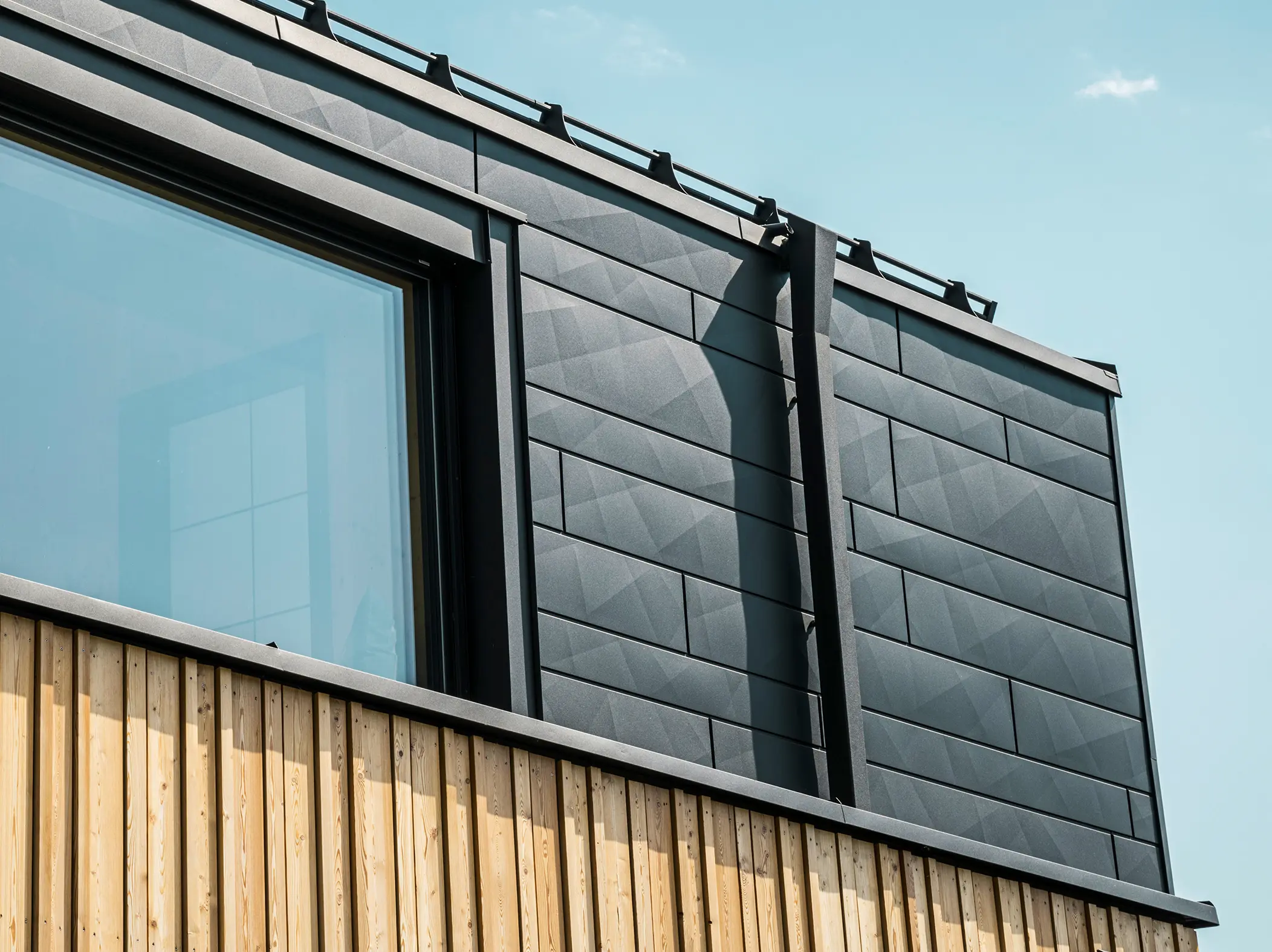 Nahaufnahme des Obergeschosses des Musterhauses Autark im Fertighauszentrum Blaue Lagune, Vösendorf (AT). Die Fassade besteht aus PREFA Siding.X Aluminiumpaneelen in P.10 Schwarz, die durch ihre spezielle Optik mit unterschiedlichen Kantungen auffallen. Darunter befindet sich eine stilvolle Holzverkleidung, die einen harmonischen Kontrast zum dunklen Aluminium bildet. Große Fenster lassen viel Licht in das Gebäudeinnere und betonen das moderne Design.