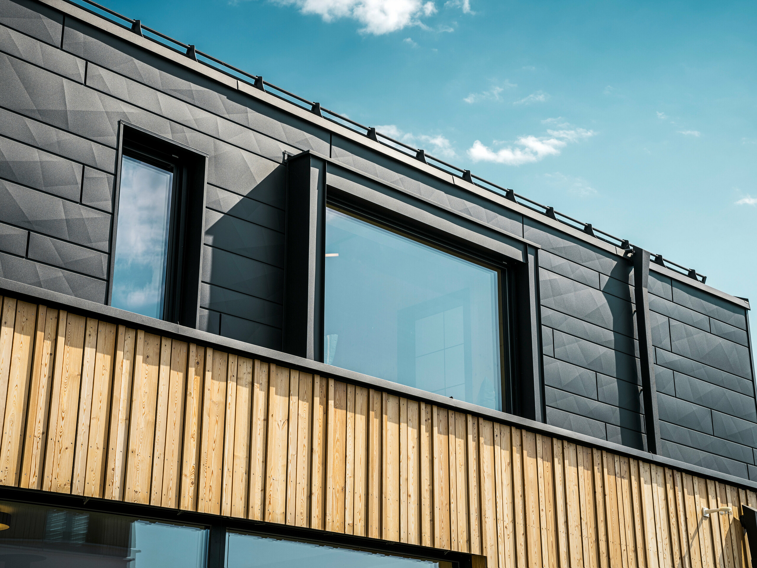 Nahaufnahme des Obergeschosses des Musterhauses Autark im Fertighauszentrum Blaue Lagune, Vösendorf (AT). Die Fassade besteht aus PREFA Siding.X Aluminiumpaneelen in P.10 Schwarz, die durch ihre spezielle Optik mit unterschiedlichen Kantungen auffallen. Darunter befindet sich eine stilvolle Holzverkleidung, die einen harmonischen Kontrast zum dunklen Aluminium bildet. Große Fenster lassen viel Licht in das Gebäudeinnere und betonen das moderne Design.
