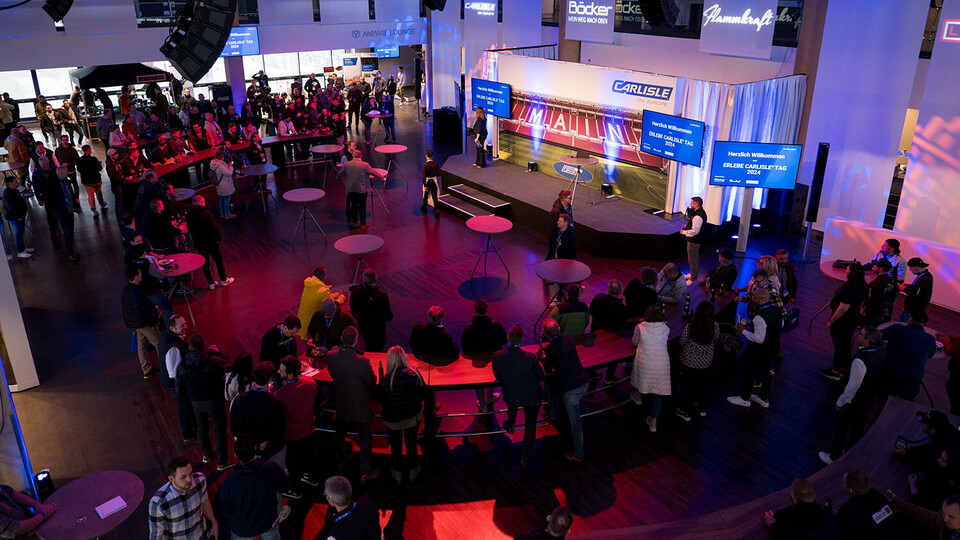 Gesamtansicht des Carlisle Branchenevents in der Mainz Arena zu sehen. Zahlreiche Besucher haben sich um die zentral platzierte Bühne versammelt, auf der Präsentationen stattfinden. Die Location ist modern ausgeleuchtet, mit großflächigem Carlisle-Branding, LED-Screens und professioneller Veranstaltungstechnik. Stehtische und Sitzbereiche fördern den Austausch der Teilnehmer, während im Hintergrund Ausstellerflächen und Partnerlogos sichtbar sind.