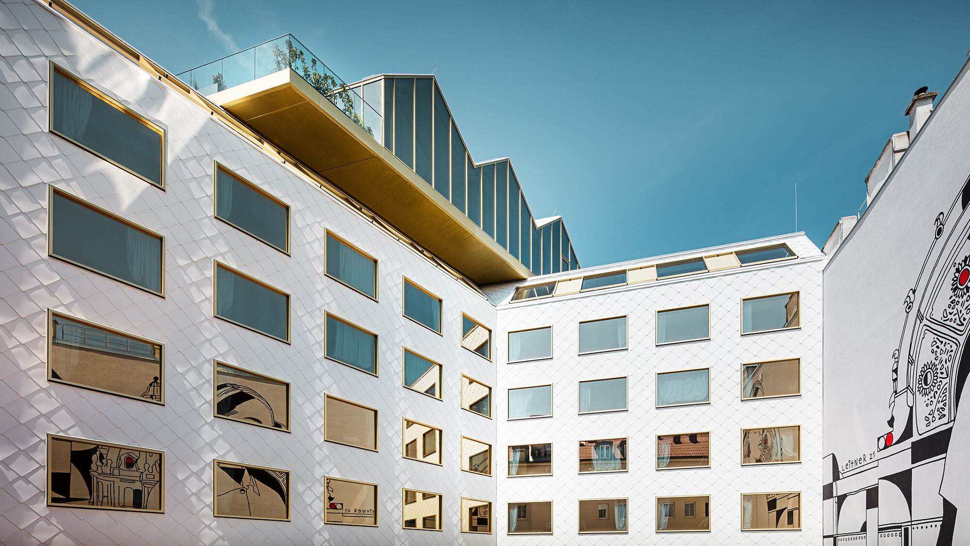 Blick auf das Hotel vom Innenhof aus; ein Wandkunstwerk spiegelt sich in den Fenstern, die Rooftop Bar kragt leicht über dem Hotel aus.