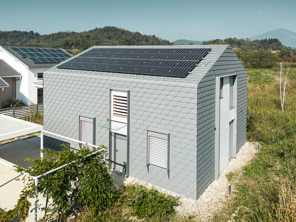 Drohnenaufnahme eines modernen Gebäudes mit einer Hülle aus PREFA Dachschindeln und Wandschindeln in der Farbe P.10 Hellgrau. Die Gebäudehülle zeichnet sich durch ein gleichmäßiges, schuppenartiges Muster aus, das eine zeitgemäße Optik bietet. Am Dach des Einfamilienhauses ist eine Photovoltaikanlage zu sehen. Im Hintergrund ist ein Haus ebenfalls mit einer Photovoltaikanlage ausgestattet wurden, was den Fokus auf Nachhaltigkeit und moderne Architektur legt.