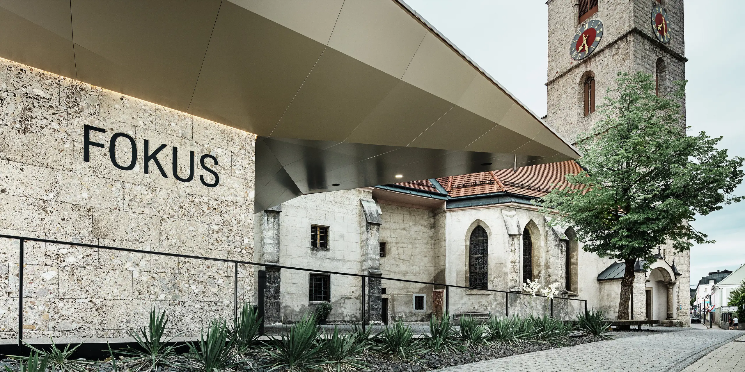 Die klare, reduzierte Geometrie des neuen Baukörpers bildet einen spannenden Kontrast zur historischen Kirche. Die glatten und langlebigen PREFA Aluminium Verbundplatten fügen sich elegant ein und sorgen für eine hochwertige, moderne und wartungsarme Fassadenlösung am Pfarrzentrum in Sierning. Die Perspektive lenkt den Blick auf das neue Foyer mit dem Schriftzug „FOKUS“.