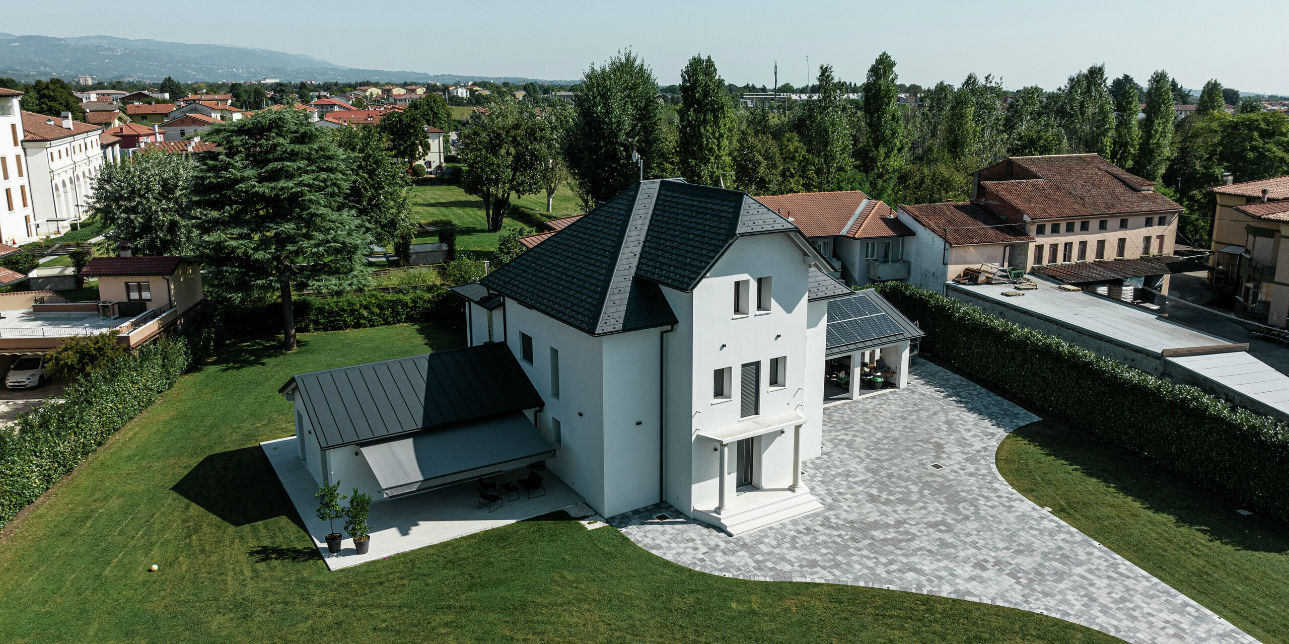 Vogelperspektive eines modernen Einfamilienhauses in Marano Vicentino, Italien, mit widerstandsfähigen PREFA Dachschindeln in P.10 Anthrazit. Die angrenzenden Nebengebäude sind mit einer Aluminium-Doppelstehfalzdeckung versehen. Die hochwertige Dachlösung aus Aluminium vereint Ästhetik, Langlebigkeit und Witterungsbeständigkeit.