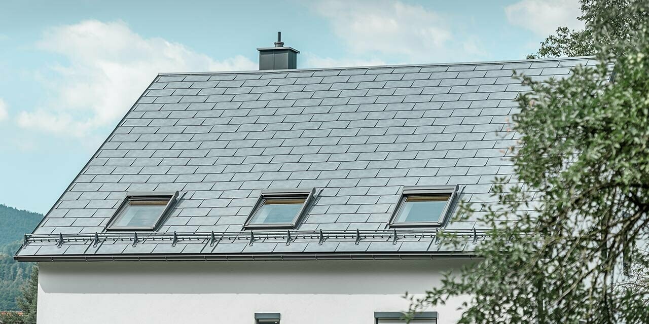 Klassisches Satteldach, eingedeckt mit der PREFA Dachplatte R.16 in Steingrau mit drei Dachfenstern und Schneerechensystem; Die Fassade ist in Weiß verputzt.