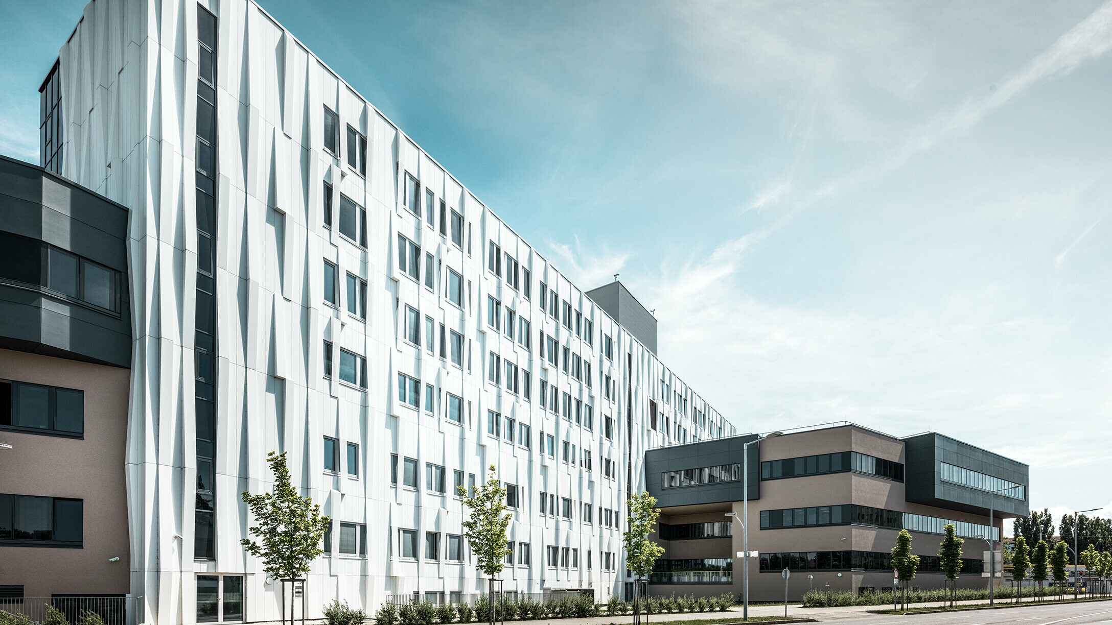 Seitliche Ansicht des Universitätsklinikums in St. Pölten, Niederösterreich. Die robuste Aluminiumfassade macht das Gebäude zu einem Blickfang in der Stadt. Im Vordergrund ist eine Straße mit einigen Bäumen zu sehen.