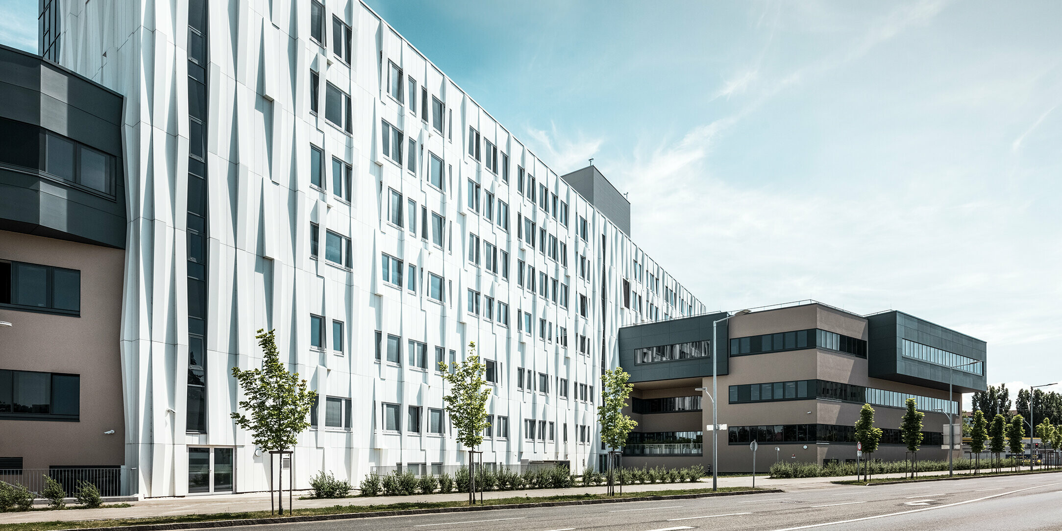 Seitliche Ansicht des Universitätsklinikums in St. Pölten, Niederösterreich. Die robuste Aluminiumfassade macht das Gebäude zu einem Blickfang in der Stadt. Im Vordergrund ist eine Straße mit einigen Bäumen zu sehen.