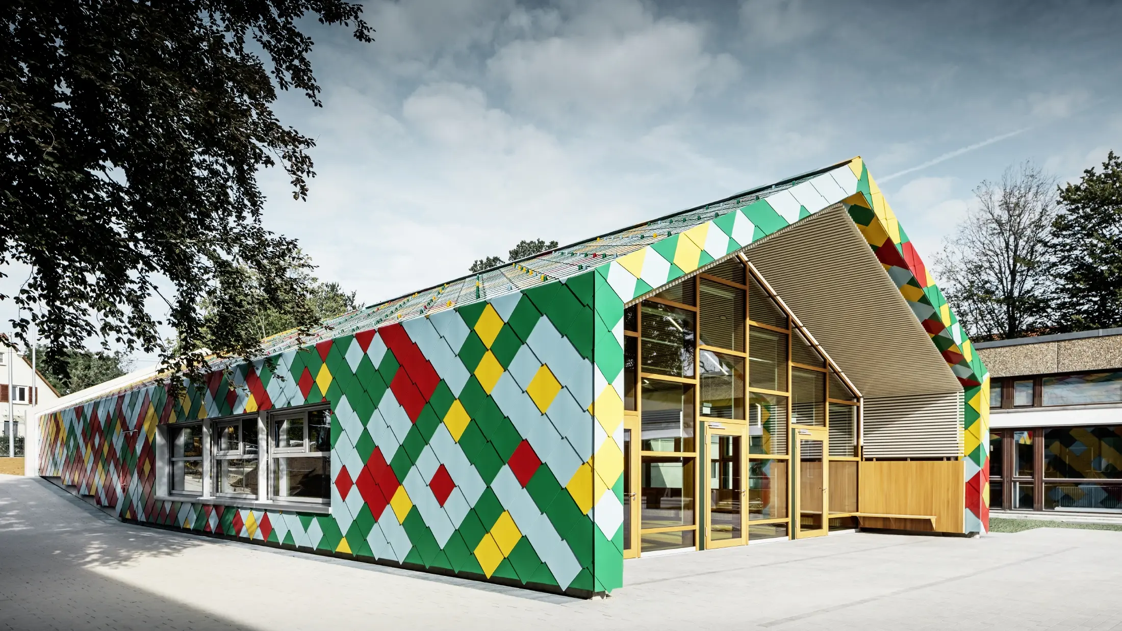 Modernes Gebäude mit auffälliger Fassade aus bunten Aluminiumrauten von PREFA in Grün, Rot, Gelb und Hellblau. Die Dach- und Wandflächen gehen fließend ineinander über und bilden ein geometrisches Muster. Der Eingangsbereich besteht aus großen Glasfronten mit Holzrahmen. Das Gebäude steht in einer grünen Umgebung mit Bäumen und weiteren Schul- oder Verwaltungsgebäuden im Hintergrund.