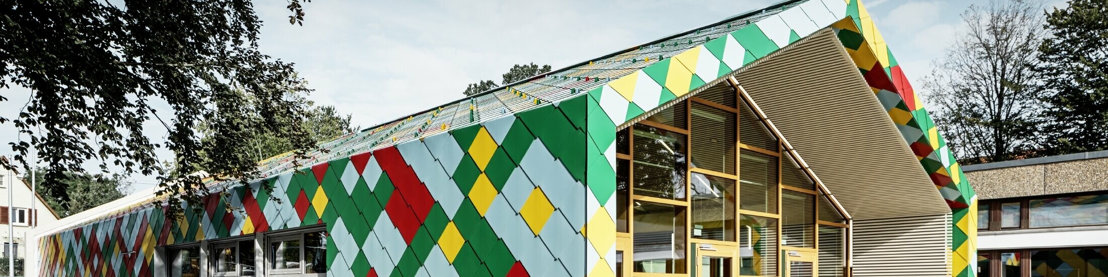 Modernes Gebäude mit auffälliger Fassade aus bunten Aluminiumrauten von PREFA in Grün, Rot, Gelb und Hellblau. Die Dach- und Wandflächen gehen fließend ineinander über und bilden ein geometrisches Muster. Der Eingangsbereich besteht aus großen Glasfronten mit Holzrahmen. Das Gebäude steht in einer grünen Umgebung mit Bäumen und weiteren Schul- oder Verwaltungsgebäuden im Hintergrund.