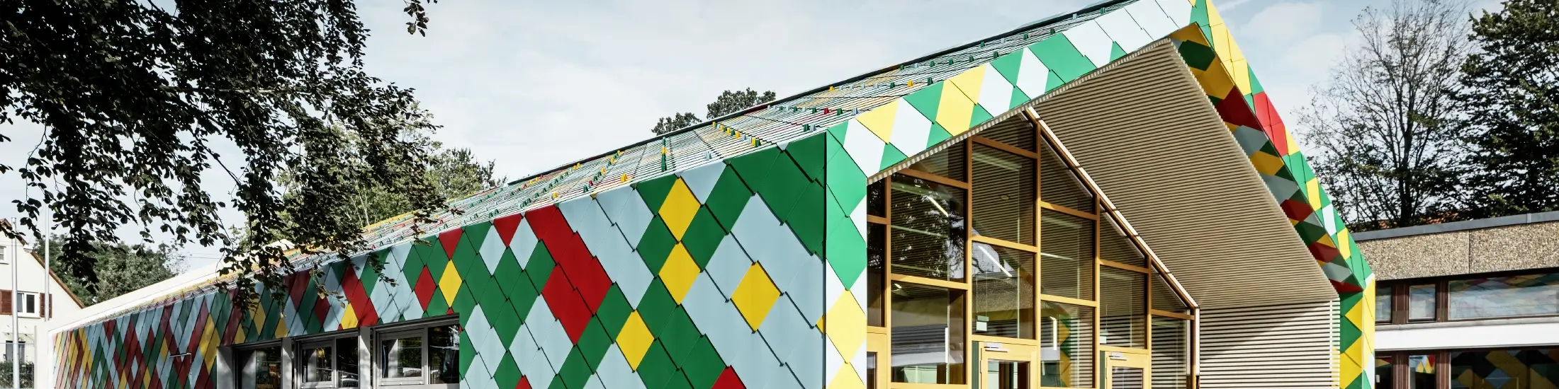 Modernes Gebäude mit auffälliger Fassade aus bunten Aluminiumrauten von PREFA in Grün, Rot, Gelb und Hellblau. Die Dach- und Wandflächen gehen fließend ineinander über und bilden ein geometrisches Muster. Der Eingangsbereich besteht aus großen Glasfronten mit Holzrahmen. Das Gebäude steht in einer grünen Umgebung mit Bäumen und weiteren Schul- oder Verwaltungsgebäuden im Hintergrund.
